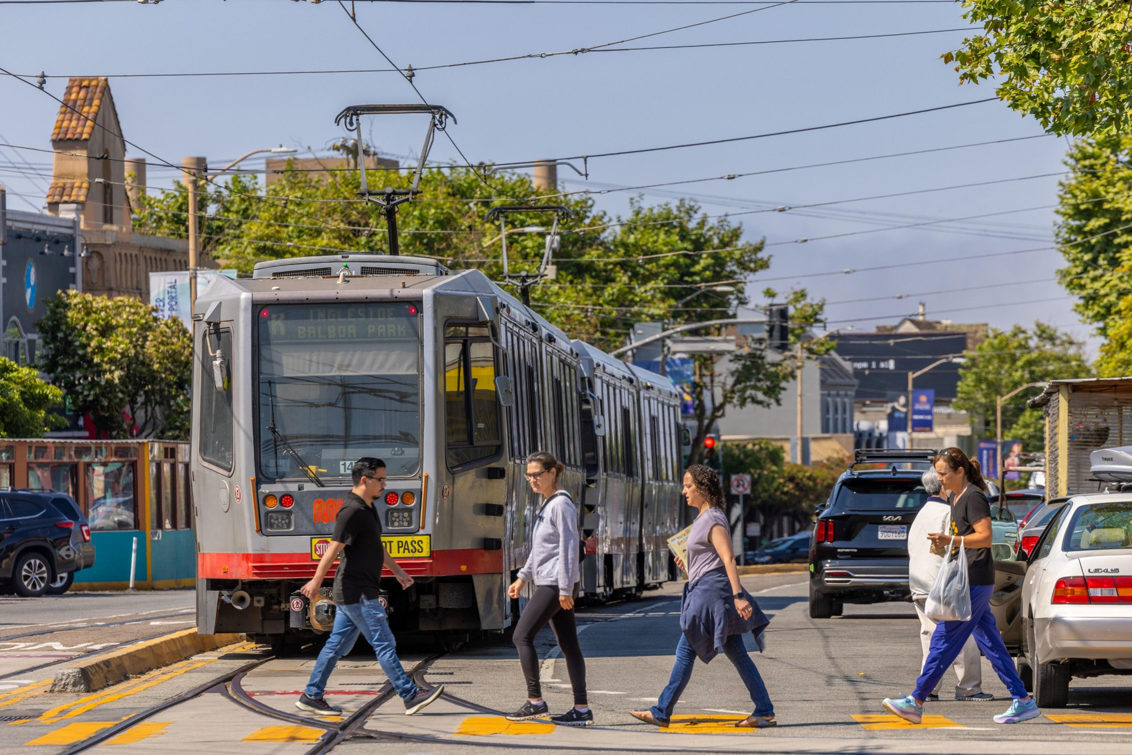 A streetcar marked "Balboa Park" awaits as five people cross the street in front of it. Vehicles line the right side of the road, with trees and buildings in the background.
