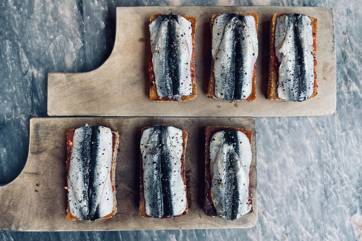 Six pieces of toasted bread each topped with a fillet of sardine and a spread, arranged on two wooden cutting boards on a marble surface.