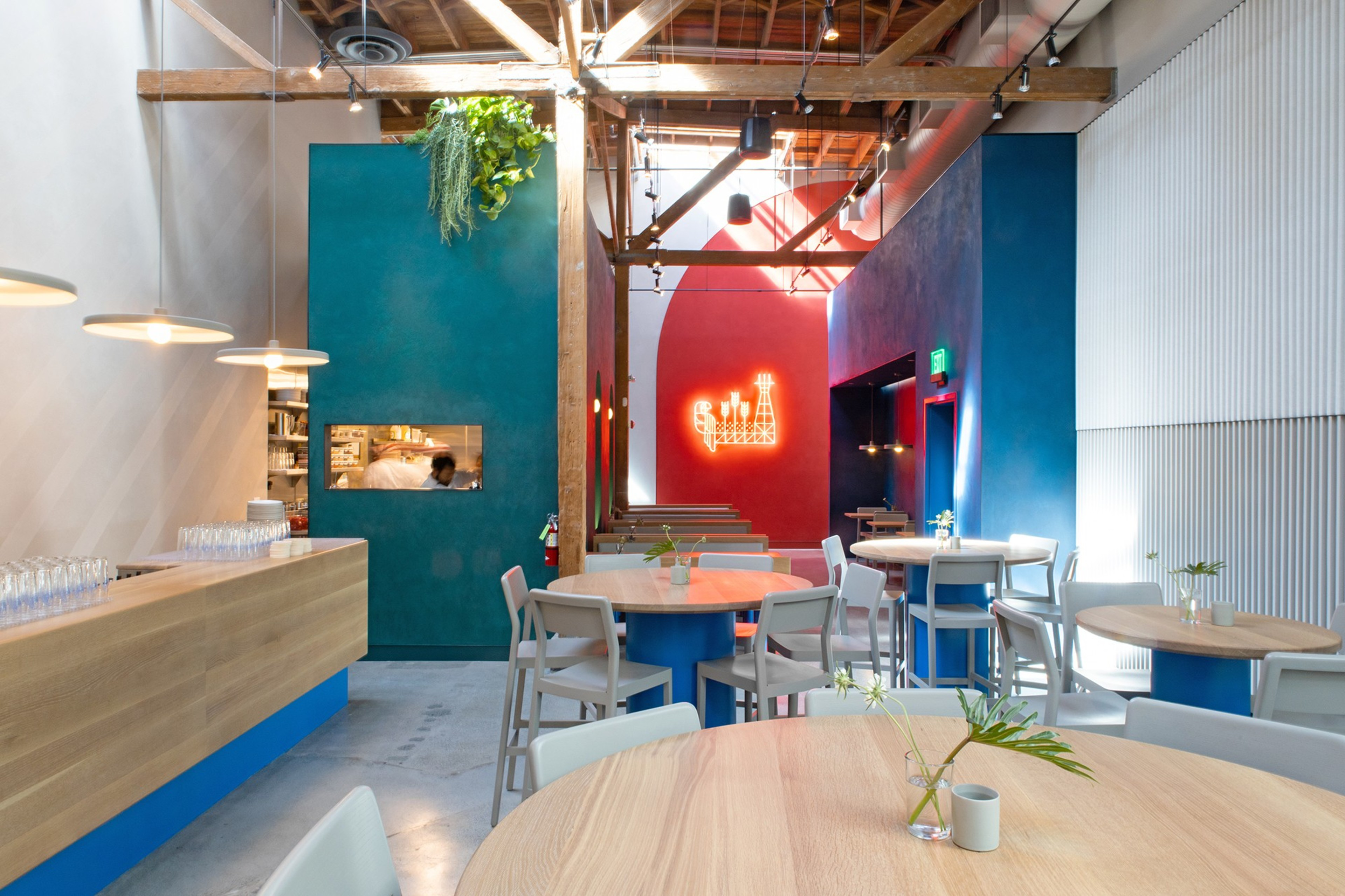 The image shows a modern restaurant with wooden tables, teal and red walls, and a neon light design. High ceilings reveal exposed beams and hanging lights.