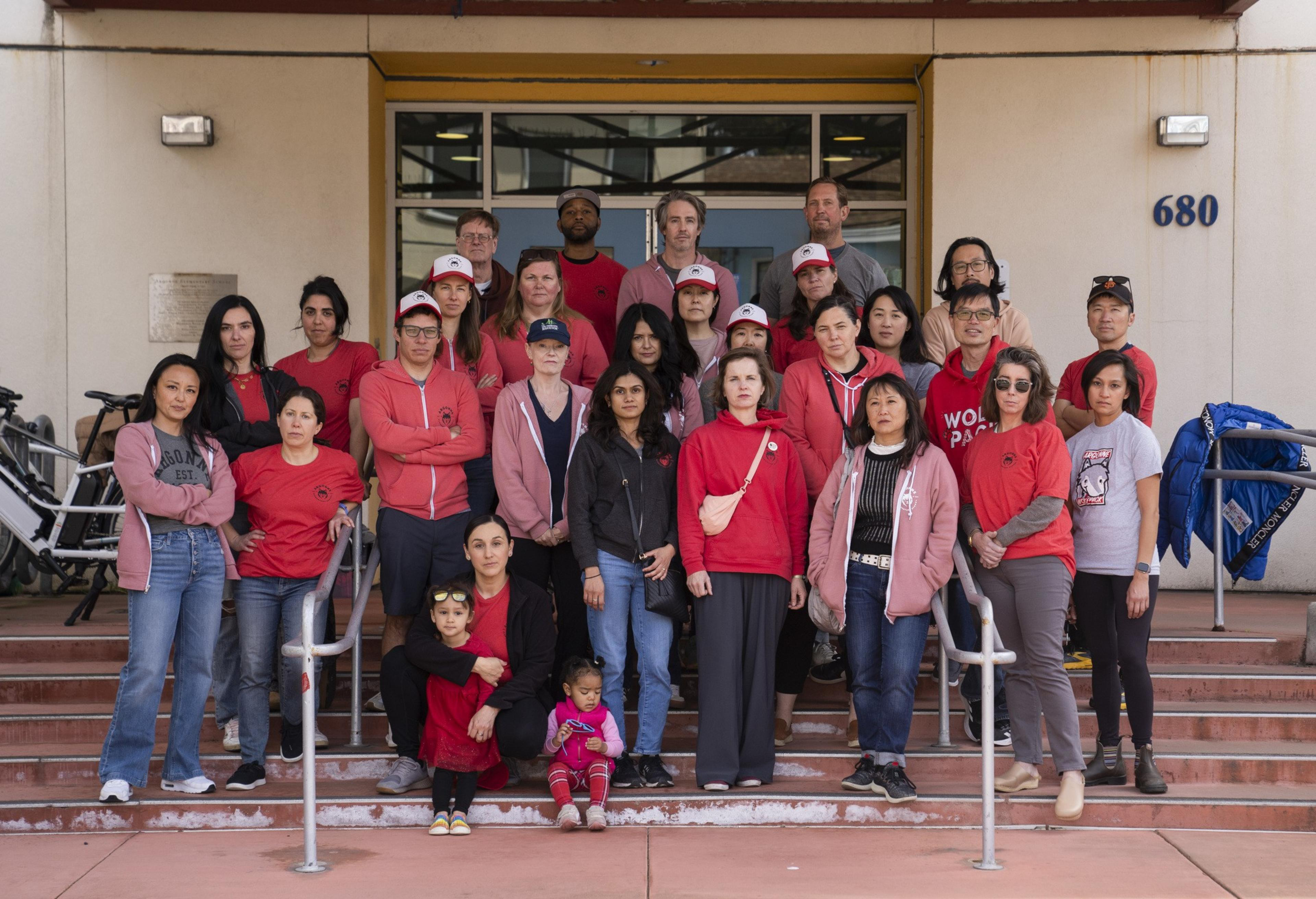 A diverse group of adults and children, some wearing red, pose on steps in front of a building numbered 680. They appear serious and united.