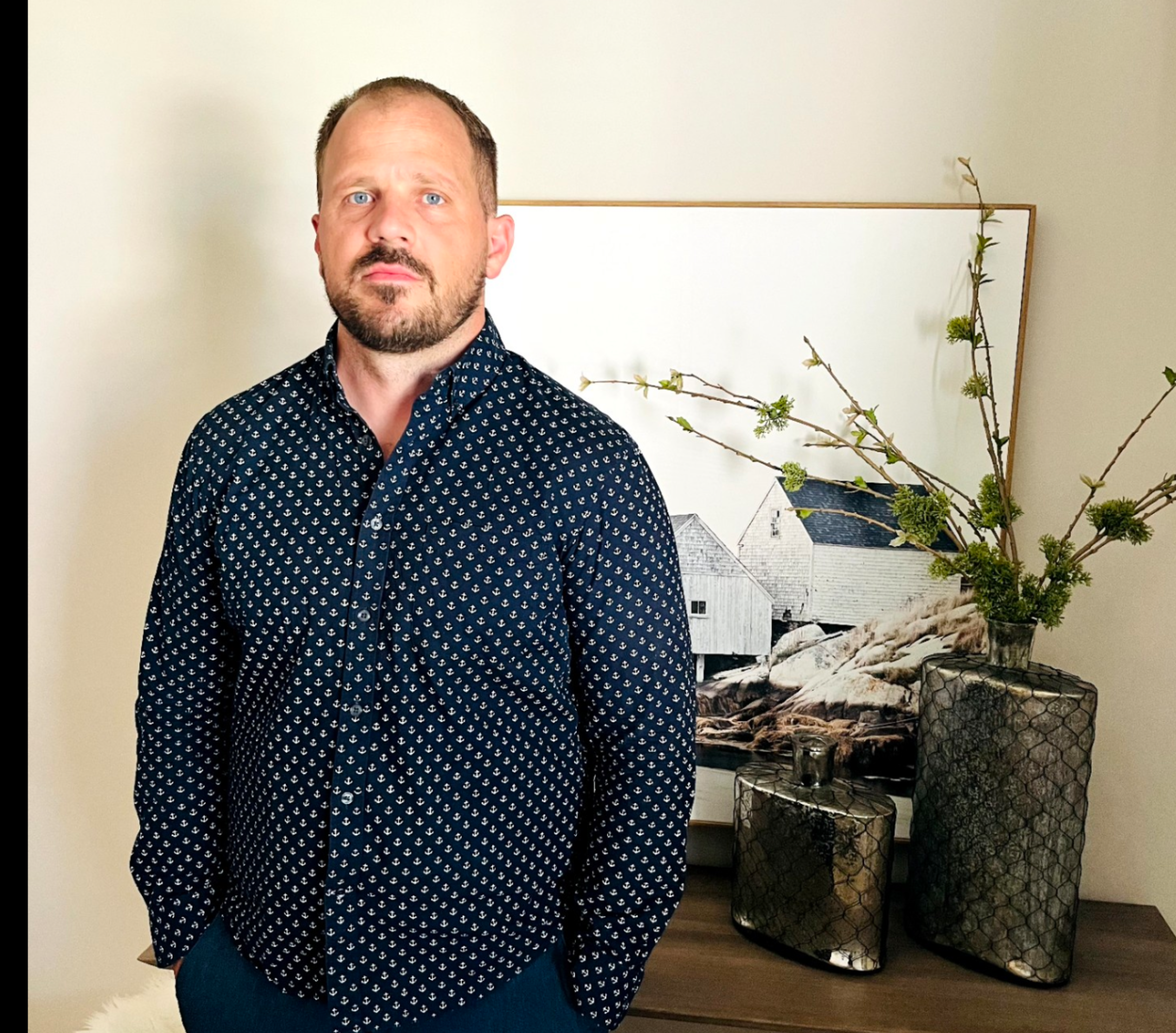 A man with a beard and blue eyes wears a navy shirt with a small white pattern, standing in front of a framed coastal house artwork and two decorative vases.