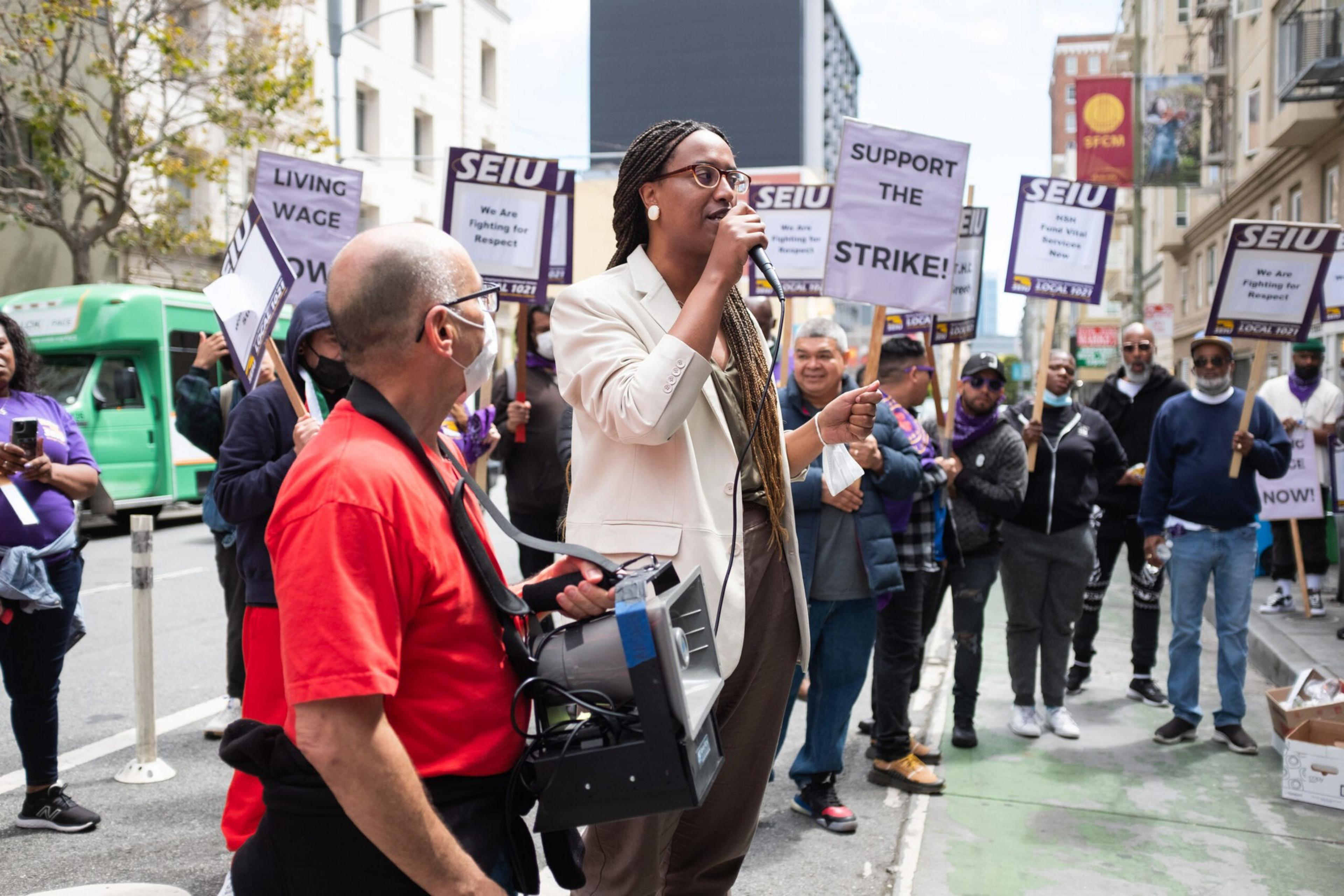 A woman speaks into a microphone while a man holds a loudspeaker; a group behind them holds signs supporting a strike and demanding a living wage.