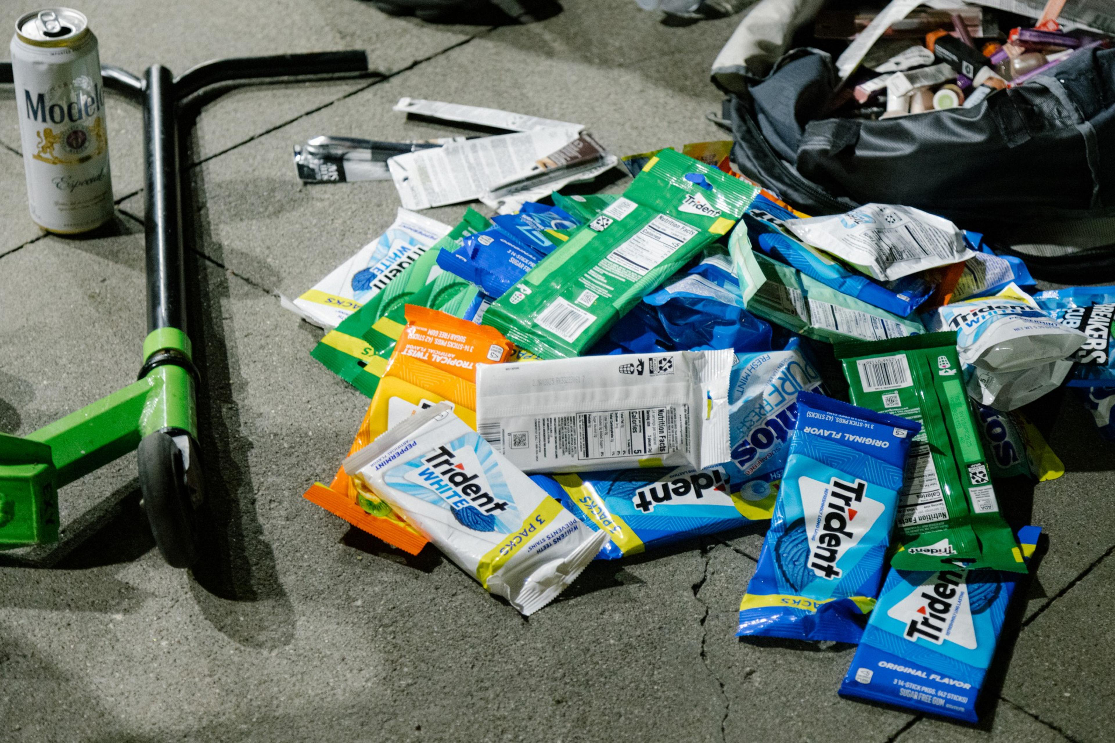 A pile of discarded chewing gum packets and a beer can, with a scooter laying nearby on a concrete surface.