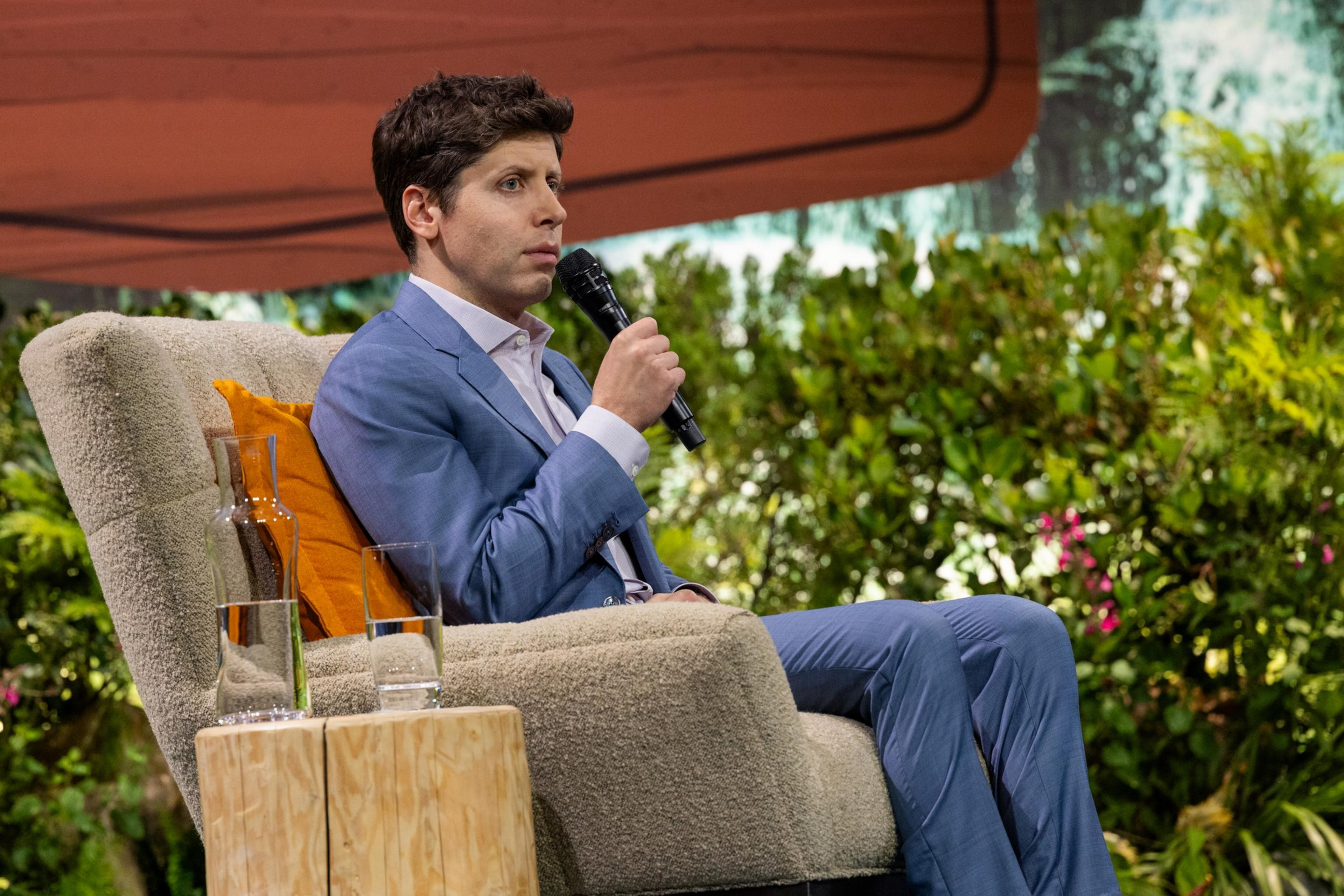 A man in a blue suit sits in a beige armchair holding a microphone, with a glass of water and a carafe on a wooden stool nearby.
