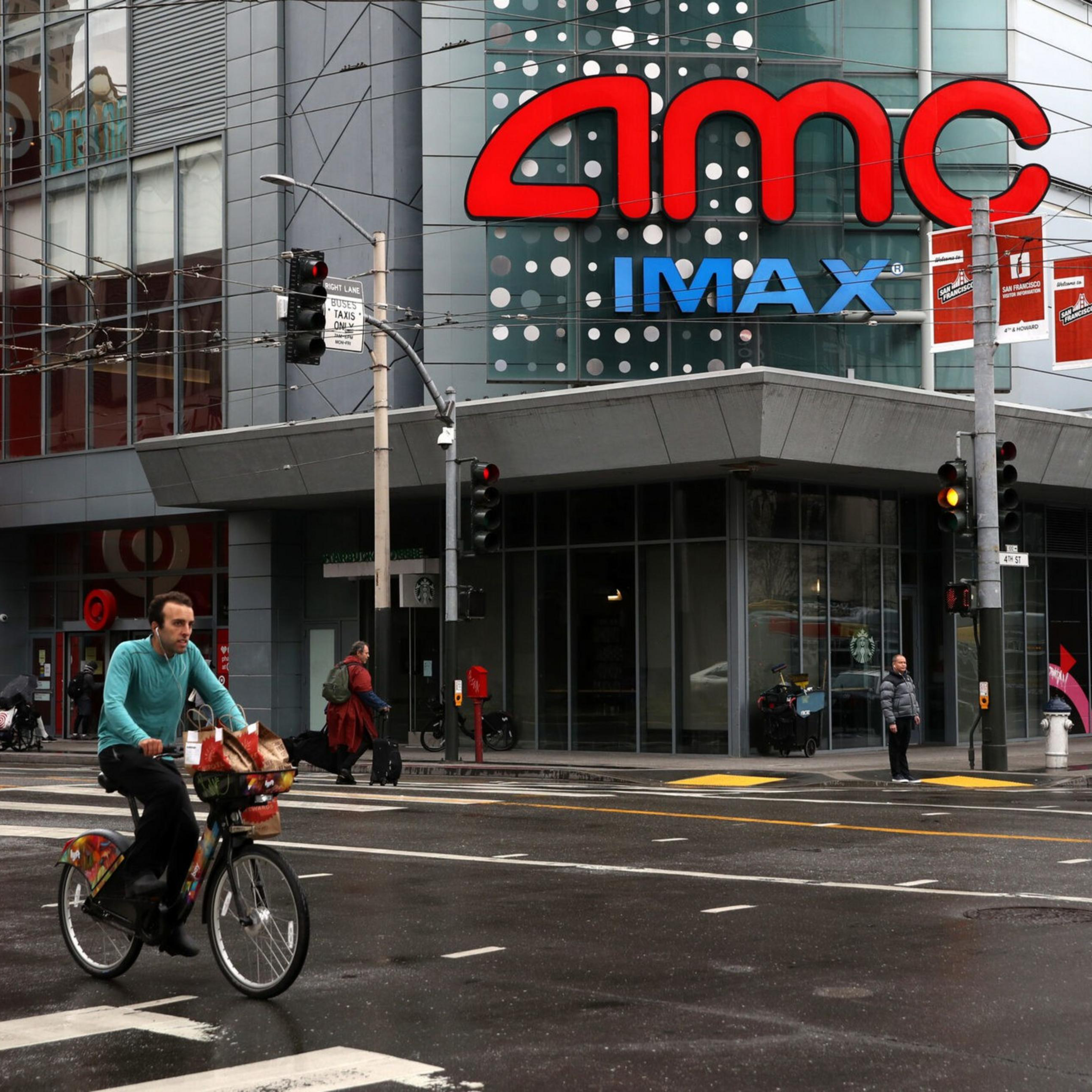 A man rides a bicycle across a wet street in front of an AMC IMAX theater and a Target store. Pedestrians walk nearby, and the traffic lights are red.