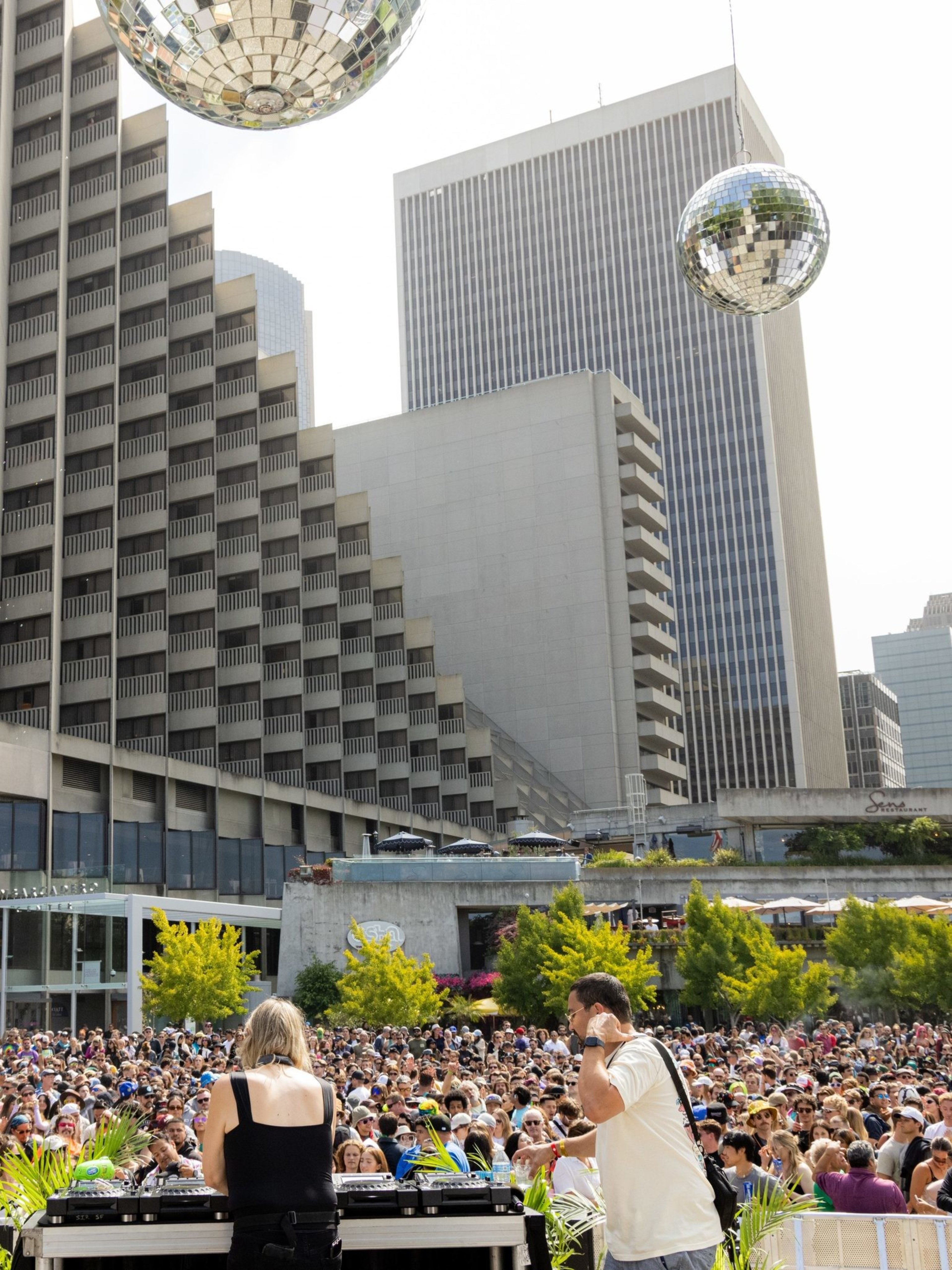 A large crowd gathers outdoors in a city plaza with towering buildings. Disco balls hang overhead, and a DJ performs on stage with lush green plants in the foreground.