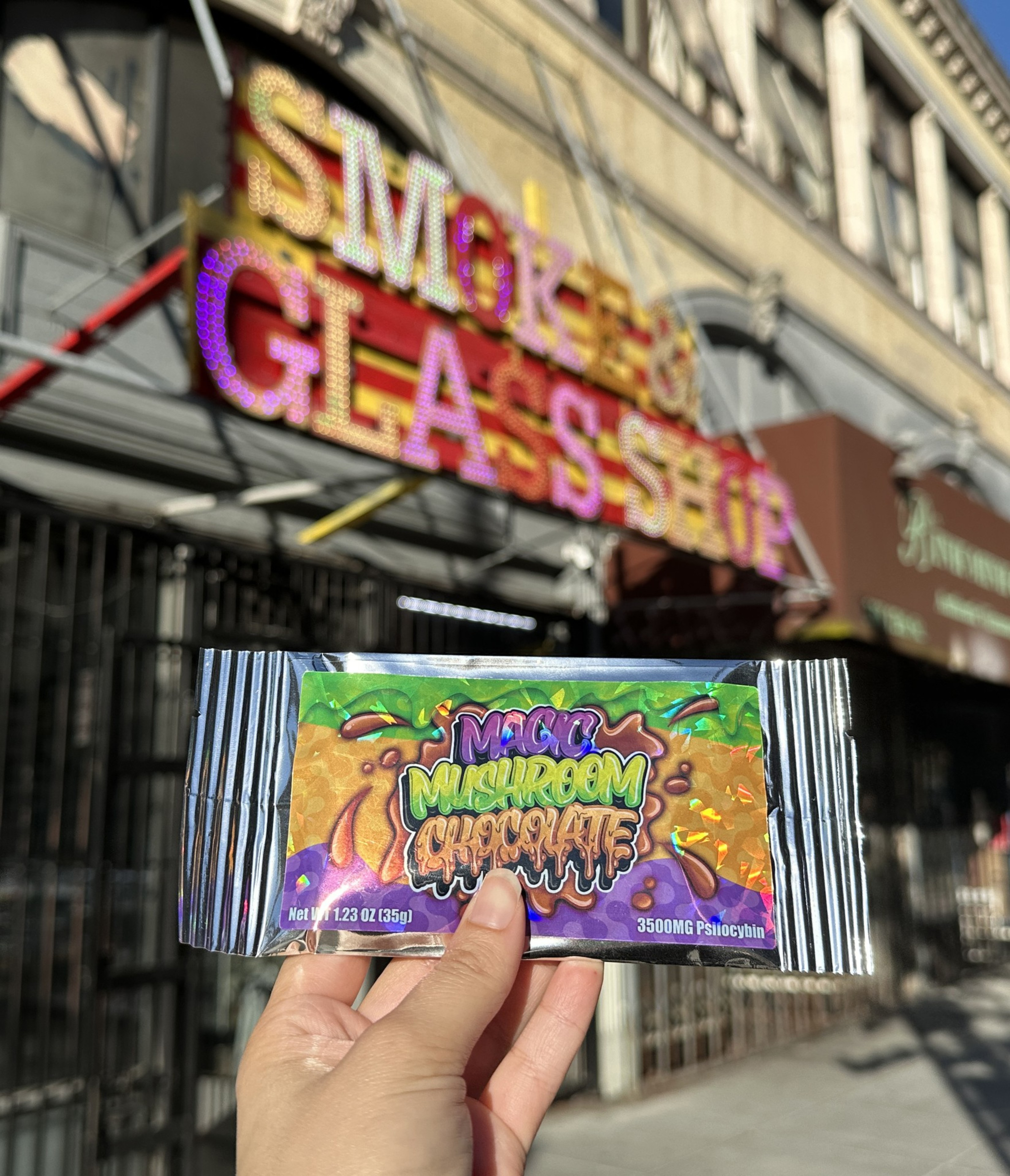 A hand holds a 'Magic Mushroom Chocolate' bar in front of a blurred Smoke and Glass shop sign.