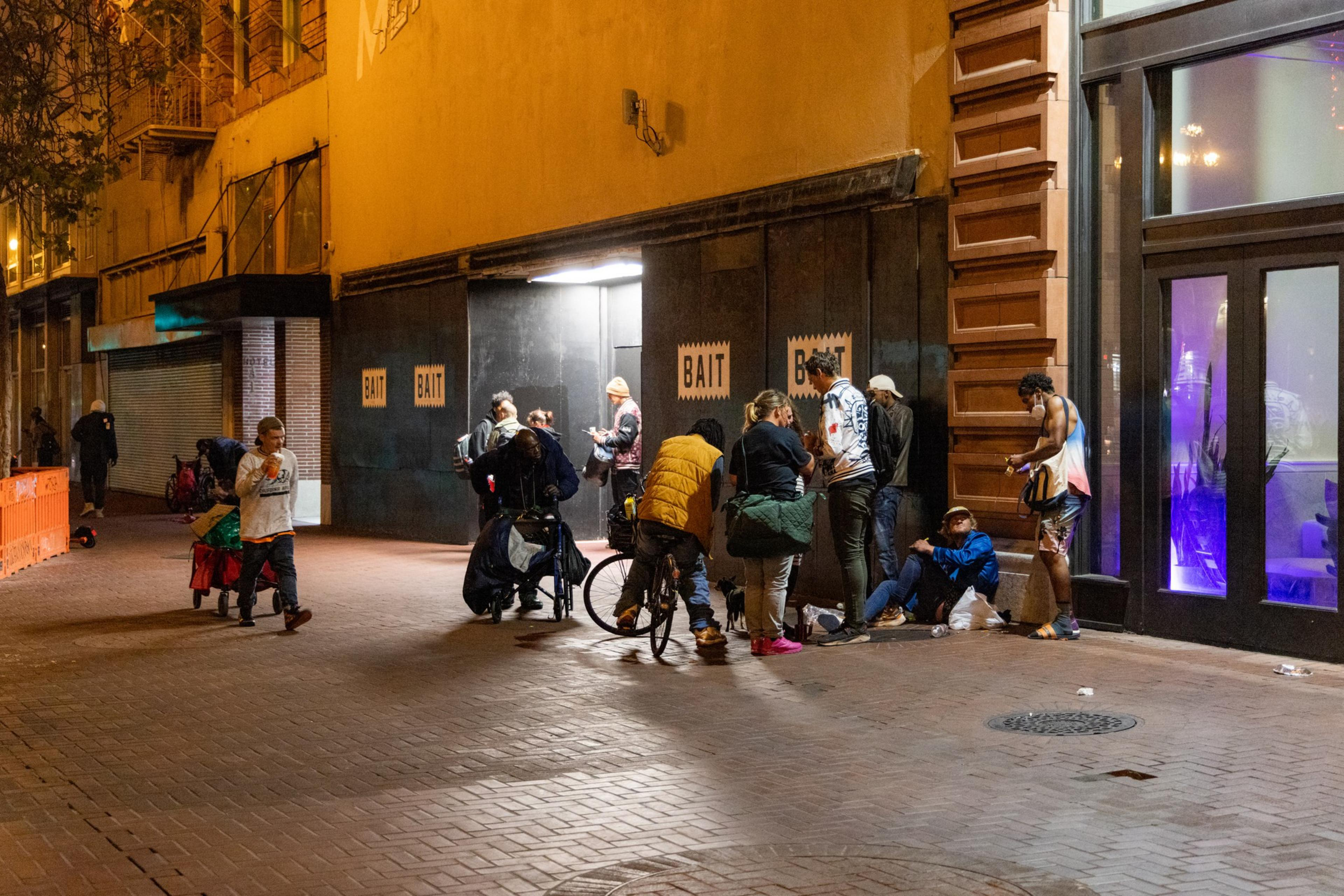 A group of people gather under dim streetlights in front of a building with signs that read &quot;BAIT.&quot; Some are on bicycles, others are sitting or standing, engaging in conversation.