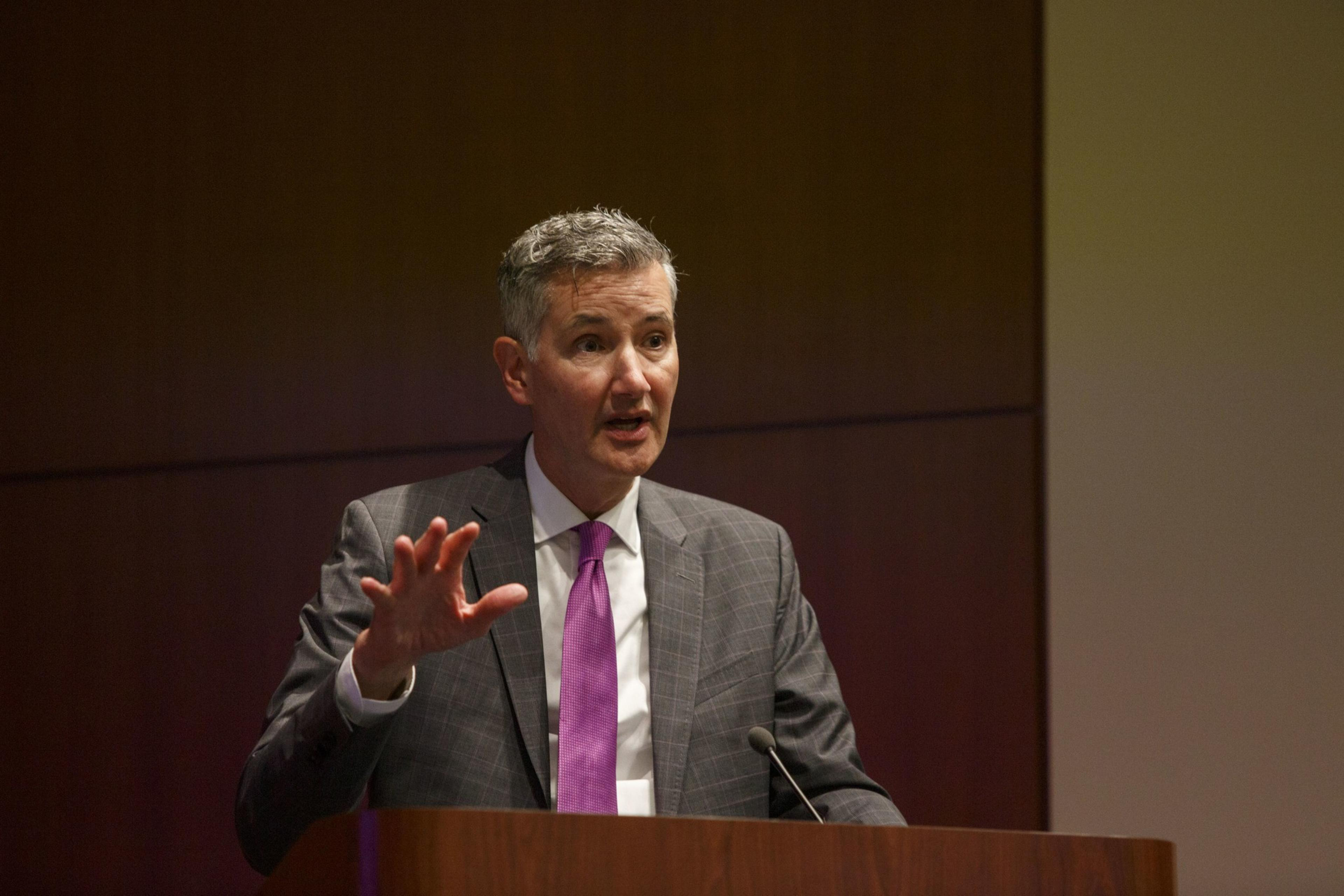 A man in a suit stands before a lecture speaking.