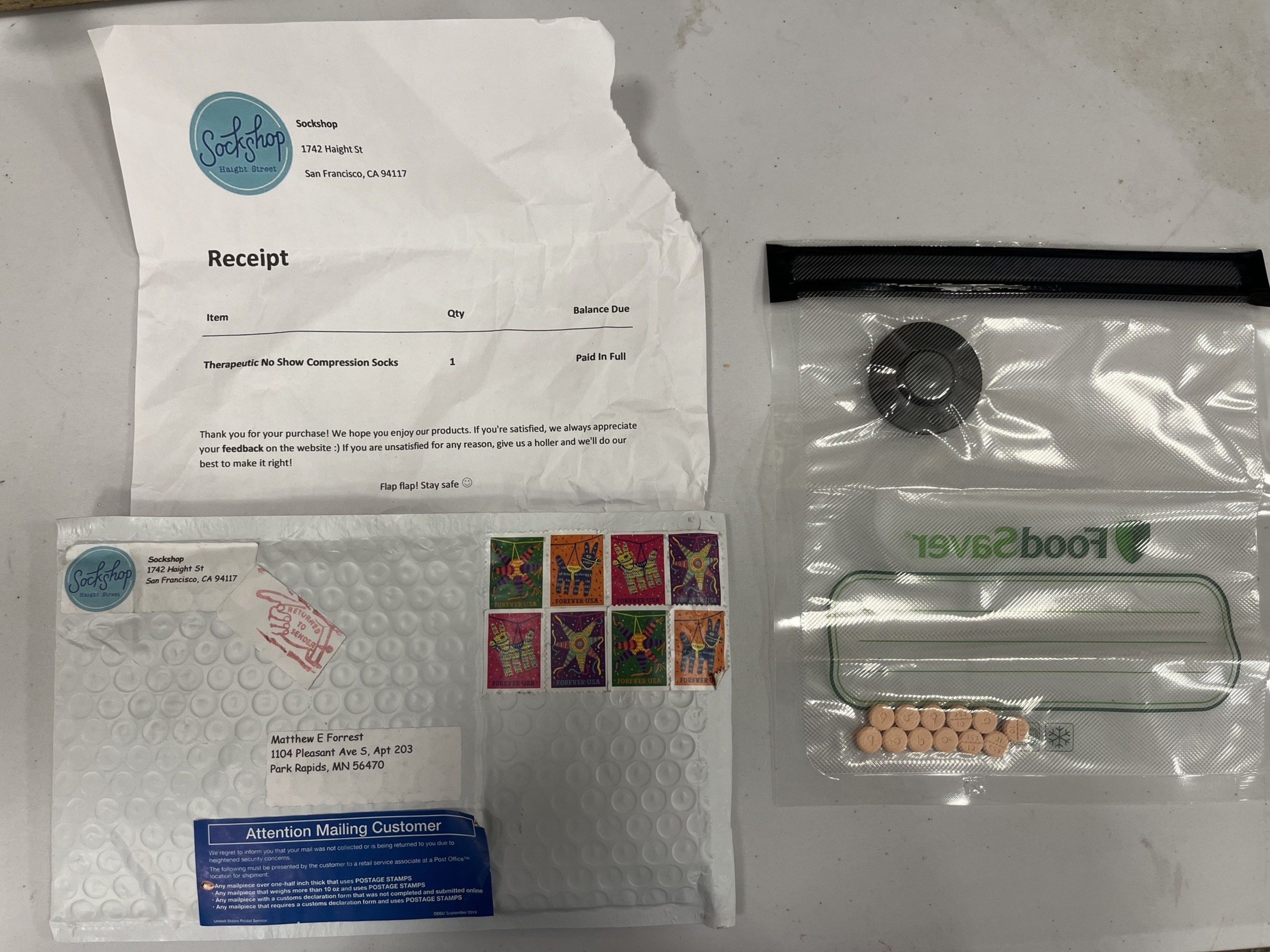 The image shows a crumpled receipt from Sockshop, a bubble mailer with stamps, and a resealable bag with a roll of black tape and small orange tablets.