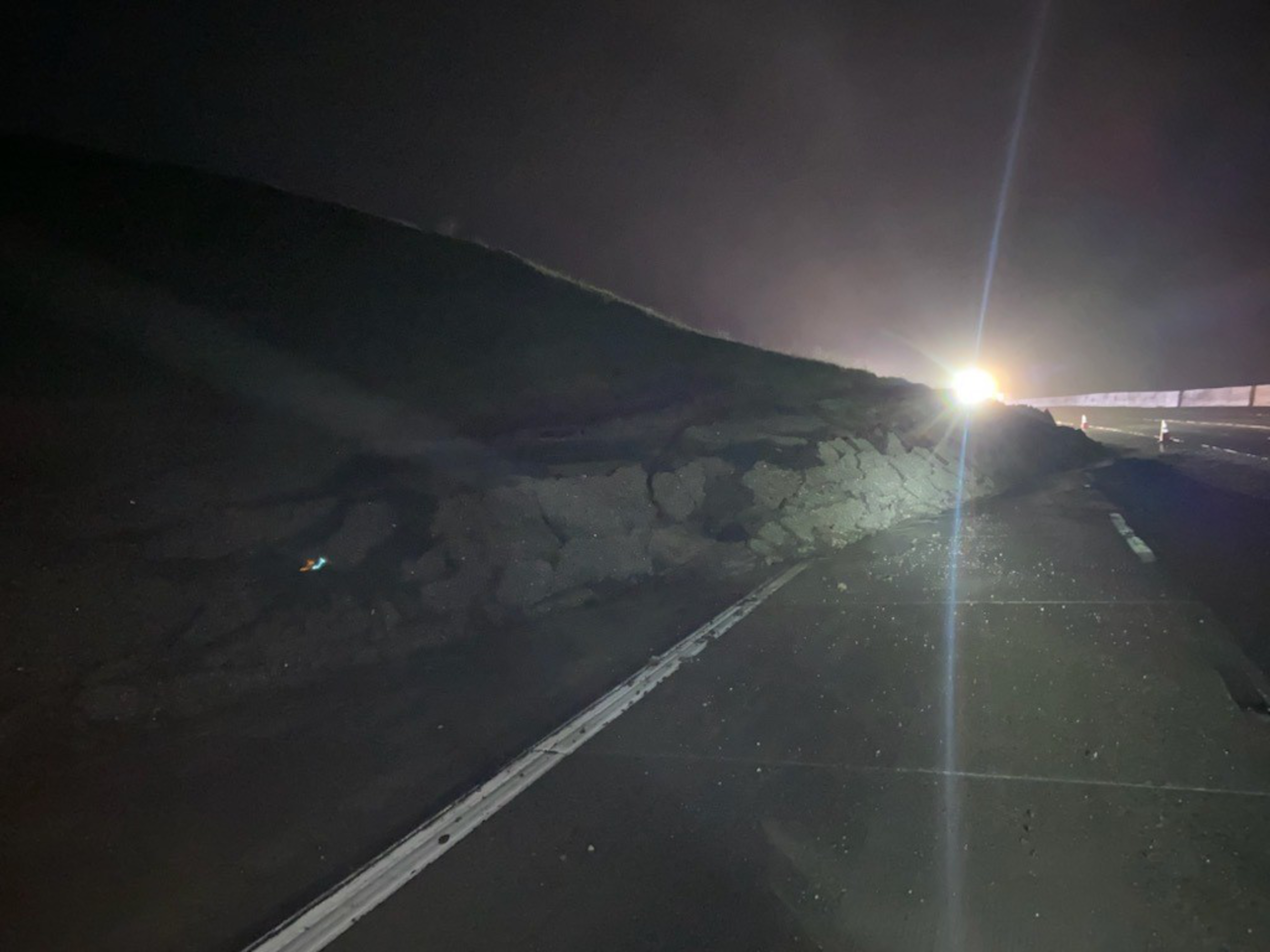 A landslide of dirt and rocks partially blocks a dark road at night, with bright headlights shining from a vehicle in the background.