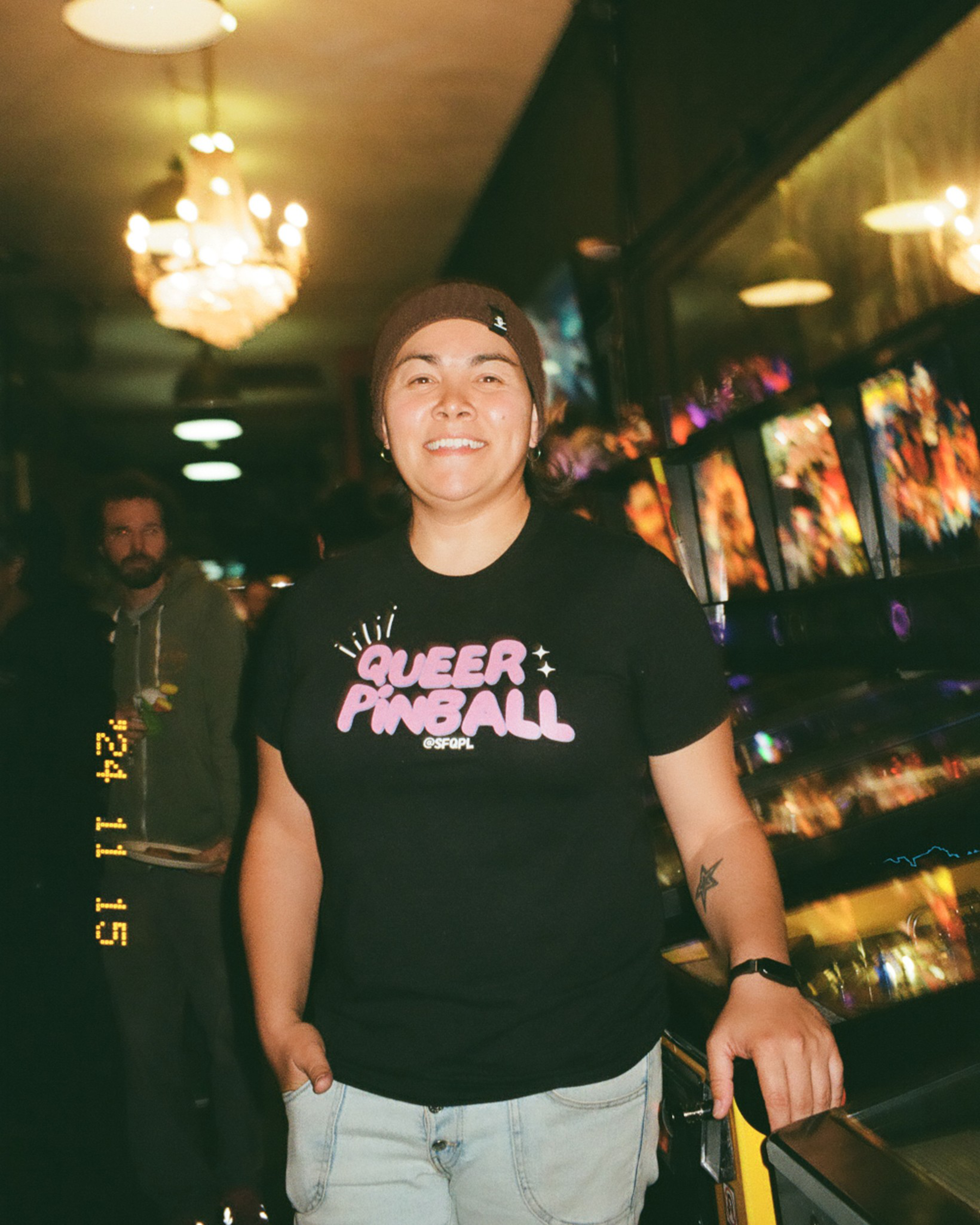 A person wearing a &quot;QUEER PINBALL&quot; t-shirt stands smiling beside a row of colorful pinball machines. The background has dim lighting with chandeliers.