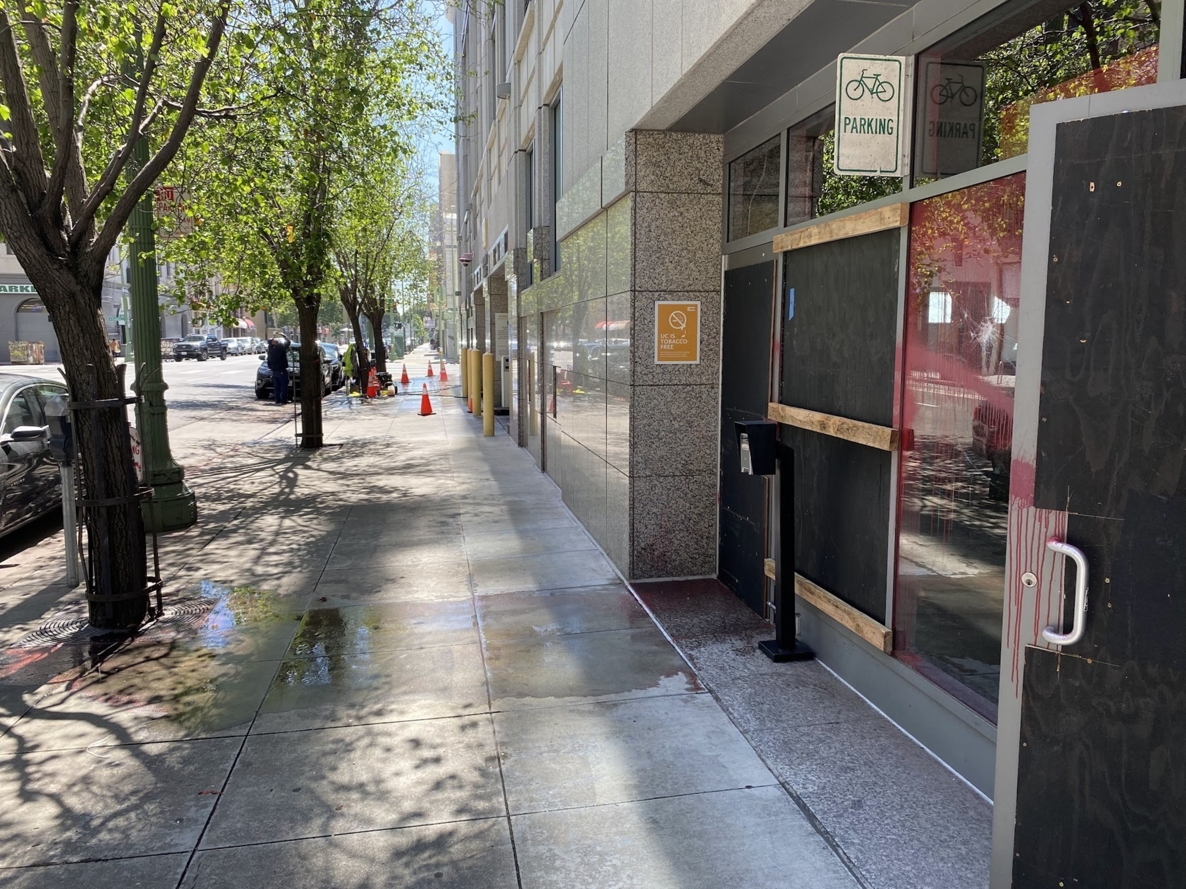 A sunlit city sidewalk, lined with green trees, leads past a building with boarded-up windows and bike parking signs.