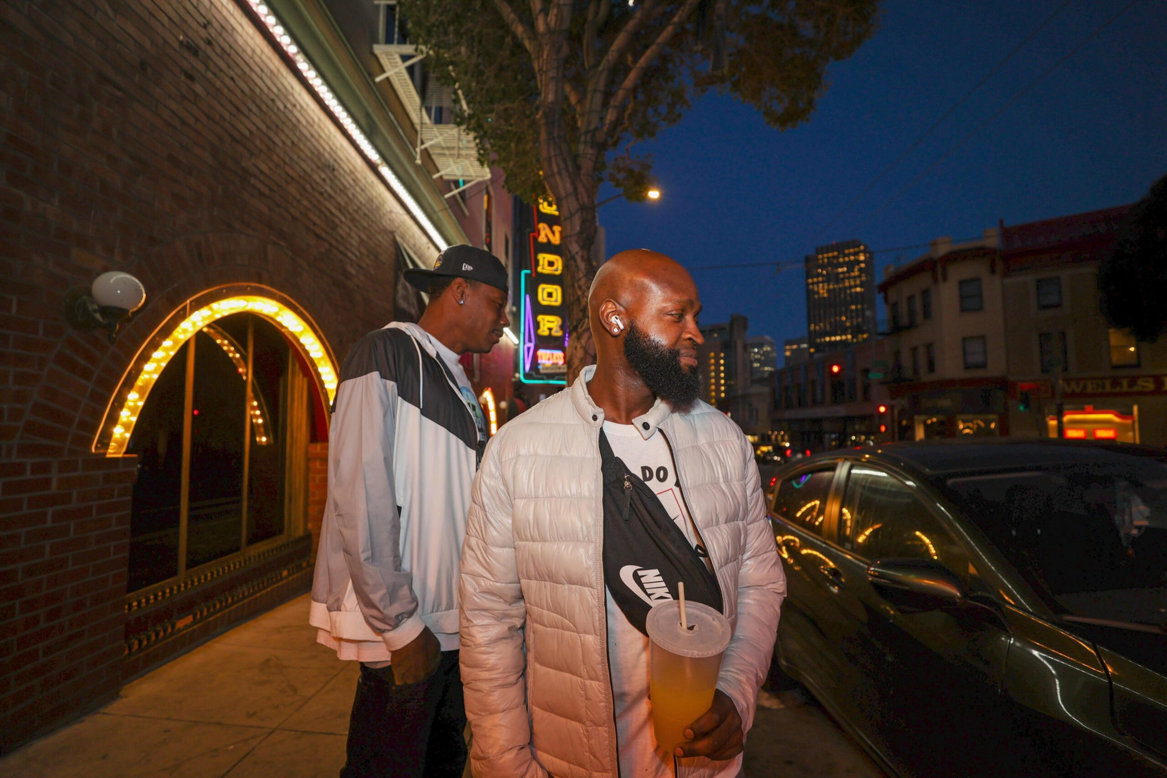 Two men stand on a city sidewalk at night; one wears a white jacket and holds a drink, while the other wears a cap and a color-block jacket near a lit building.