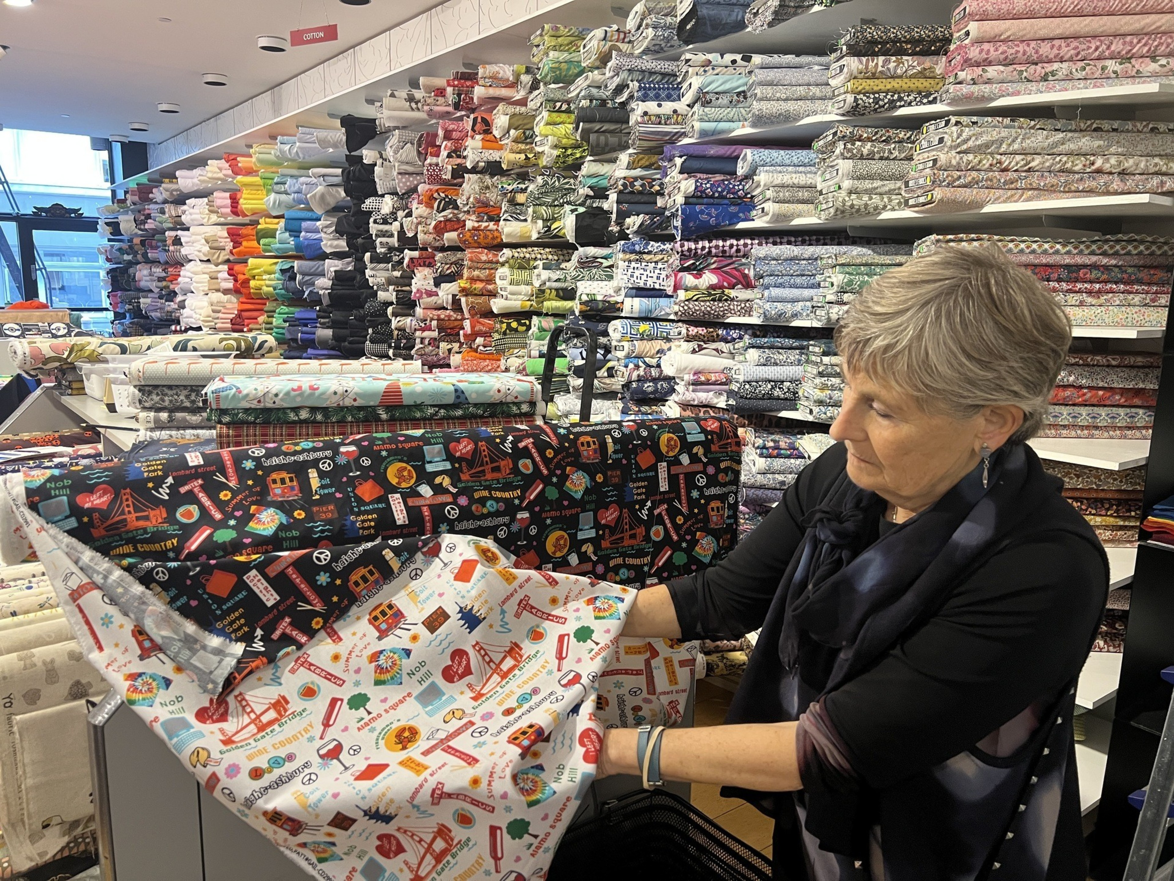A woman in a black long-sleeved shirt holds up a bolt of fabric that has images of San Francisco printed on it, with a wall of fabric behind her.