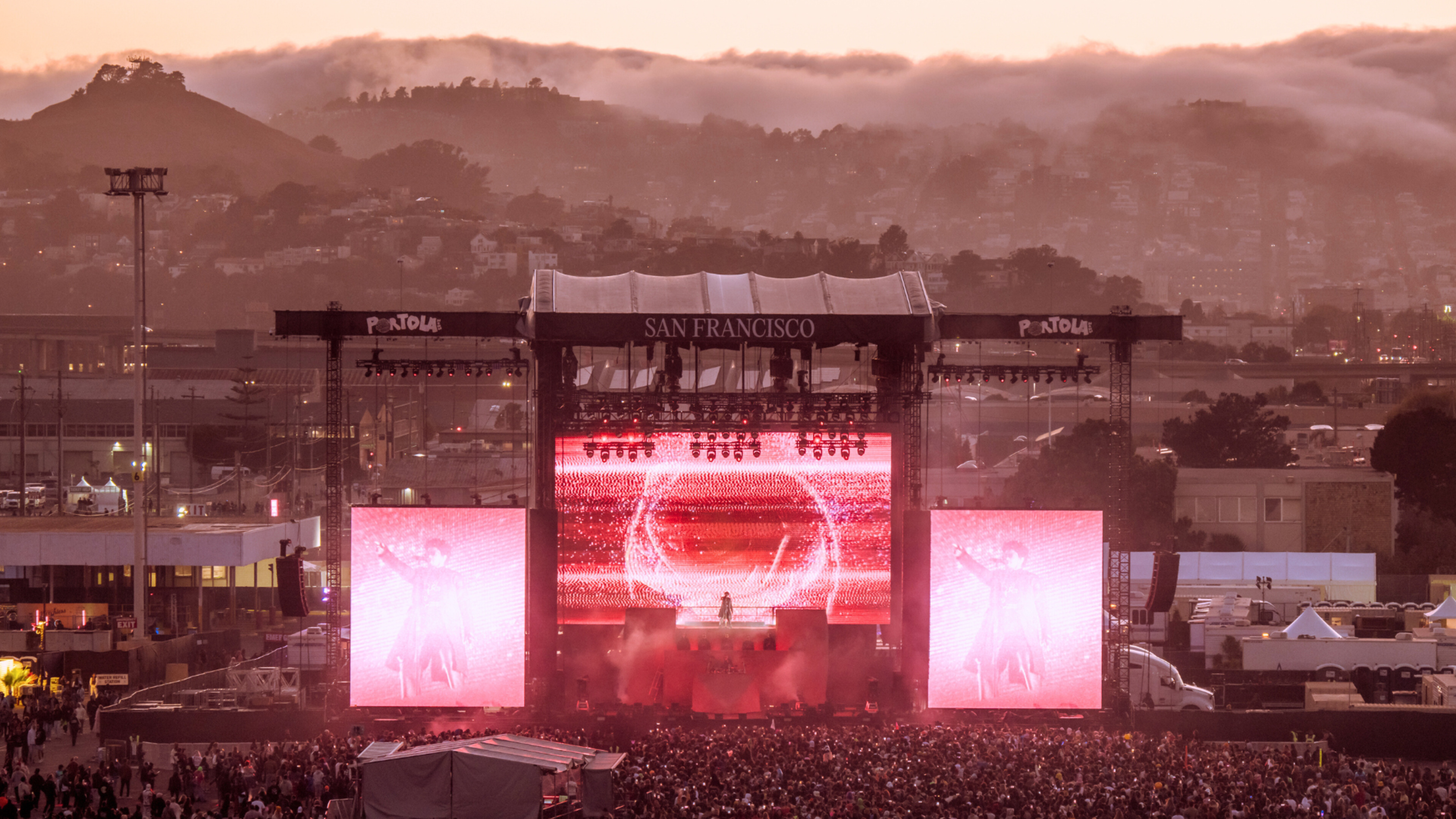 A concert stage in San Francisco with large screens showing performers, surrounded by a crowd at dusk, while fog blankets the hills in the background.