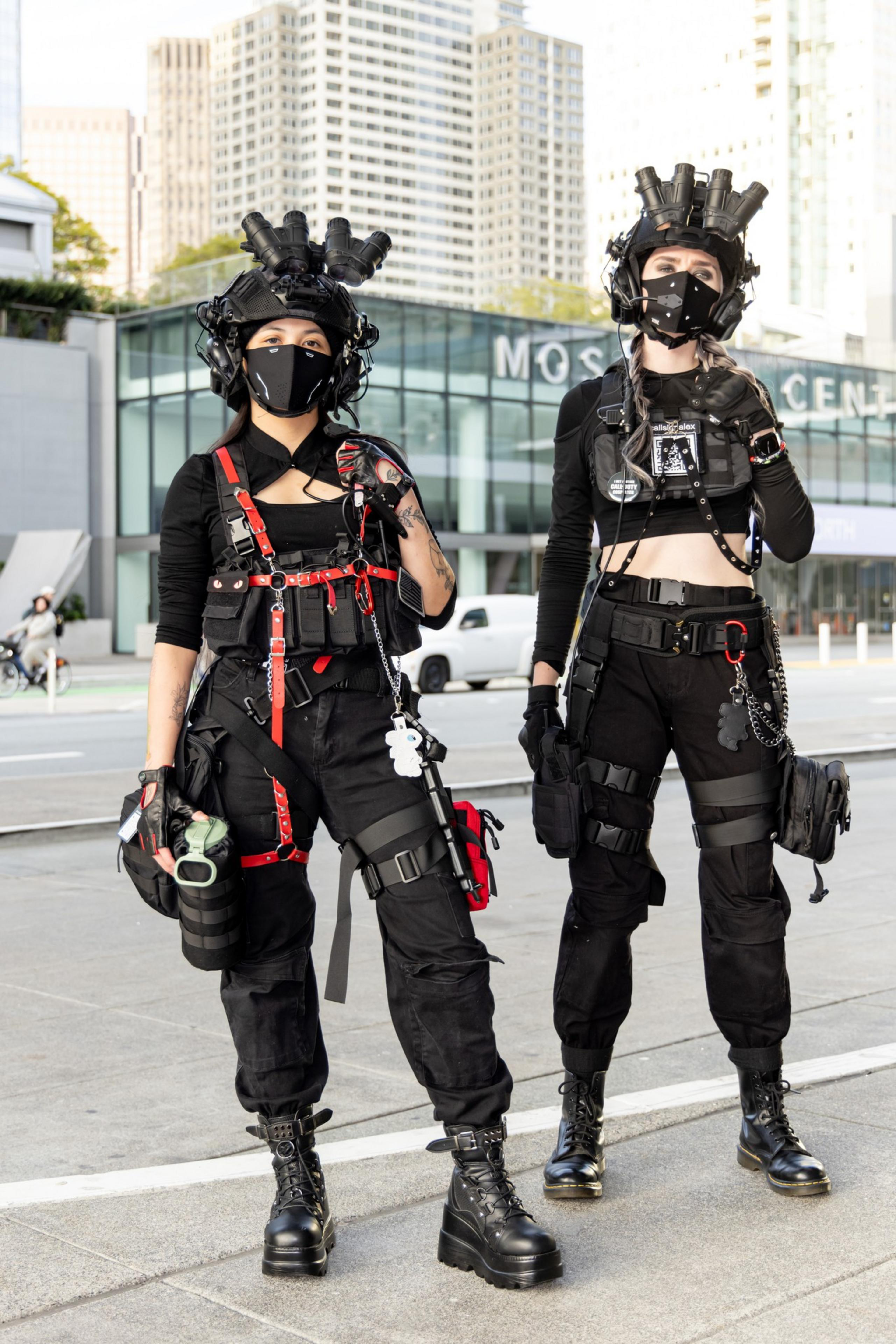 Two individuals in tactical gear and black clothing, wearing face masks and headgear with goggles, stand confidently in an urban setting.