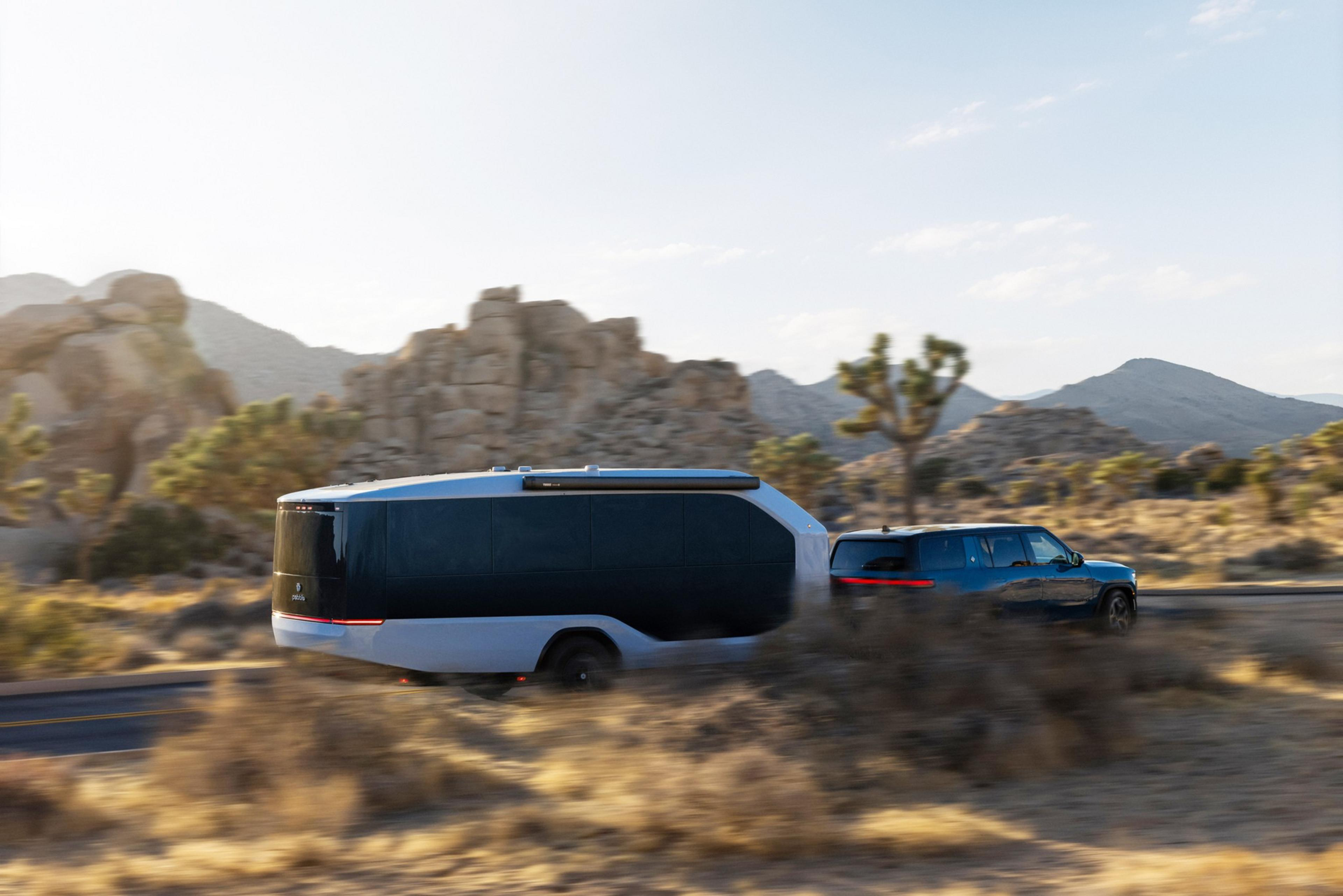 A blue SUV is towing a sleek, modern trailer through a desert landscape with rocky hills and sparse vegetation under a clear sky.