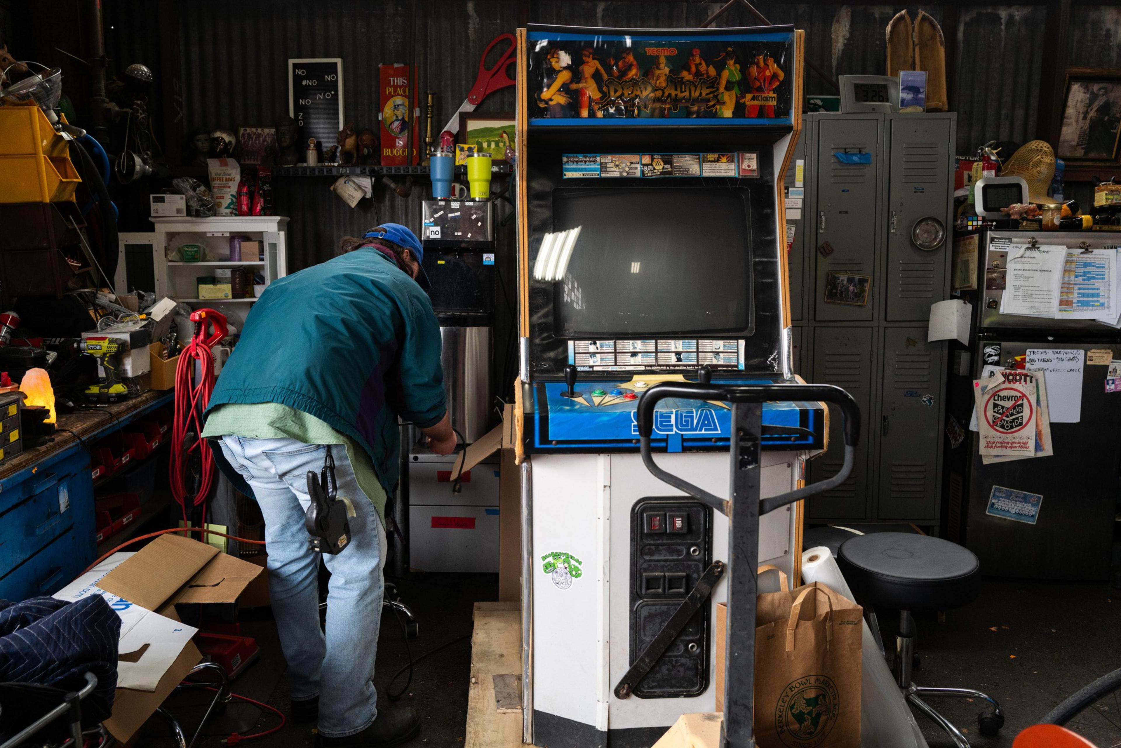 A person in a workshop bends over near a vintage arcade machine labeled &quot;Sega.&quot; The space is cluttered with tools, boxes, and lockers.