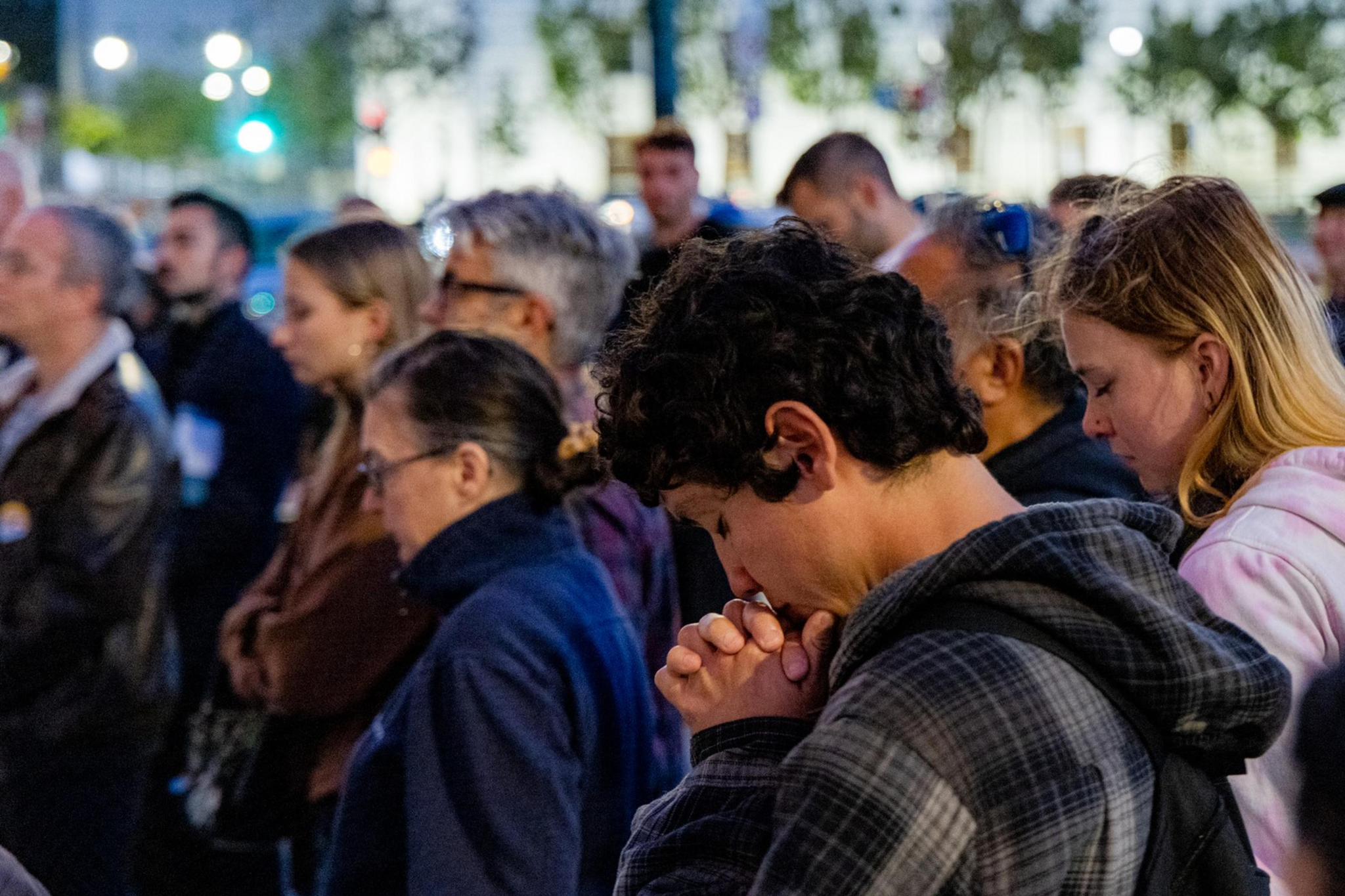 A group of people are standing close together, appearing solemn. Many have their heads bowed and eyes closed, possibly in prayer or reflection.