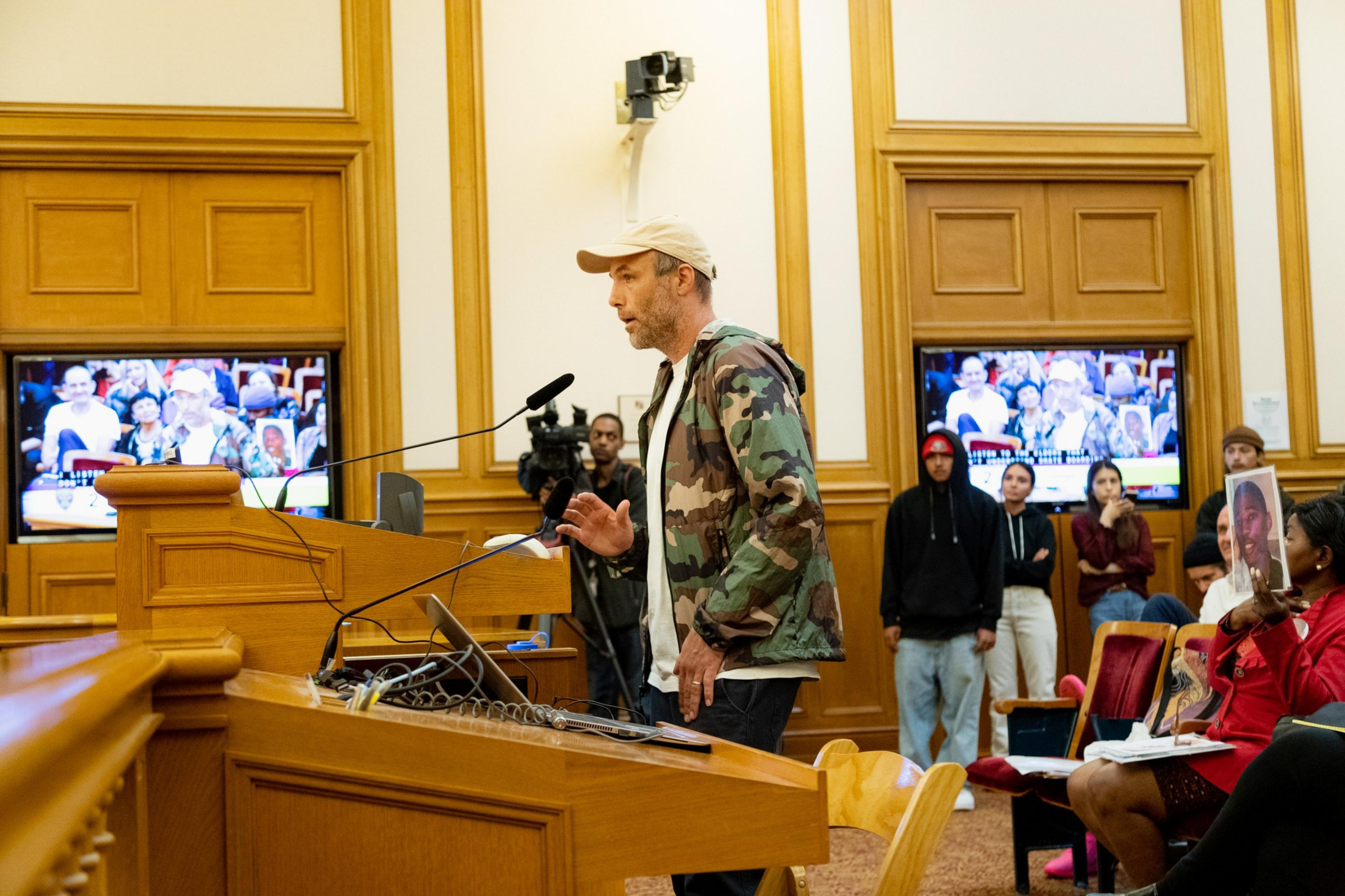 A man in a camouflage jacket and beige cap speaks at a wooden podium in a courtroom, while people watch and hold photos of individuals.