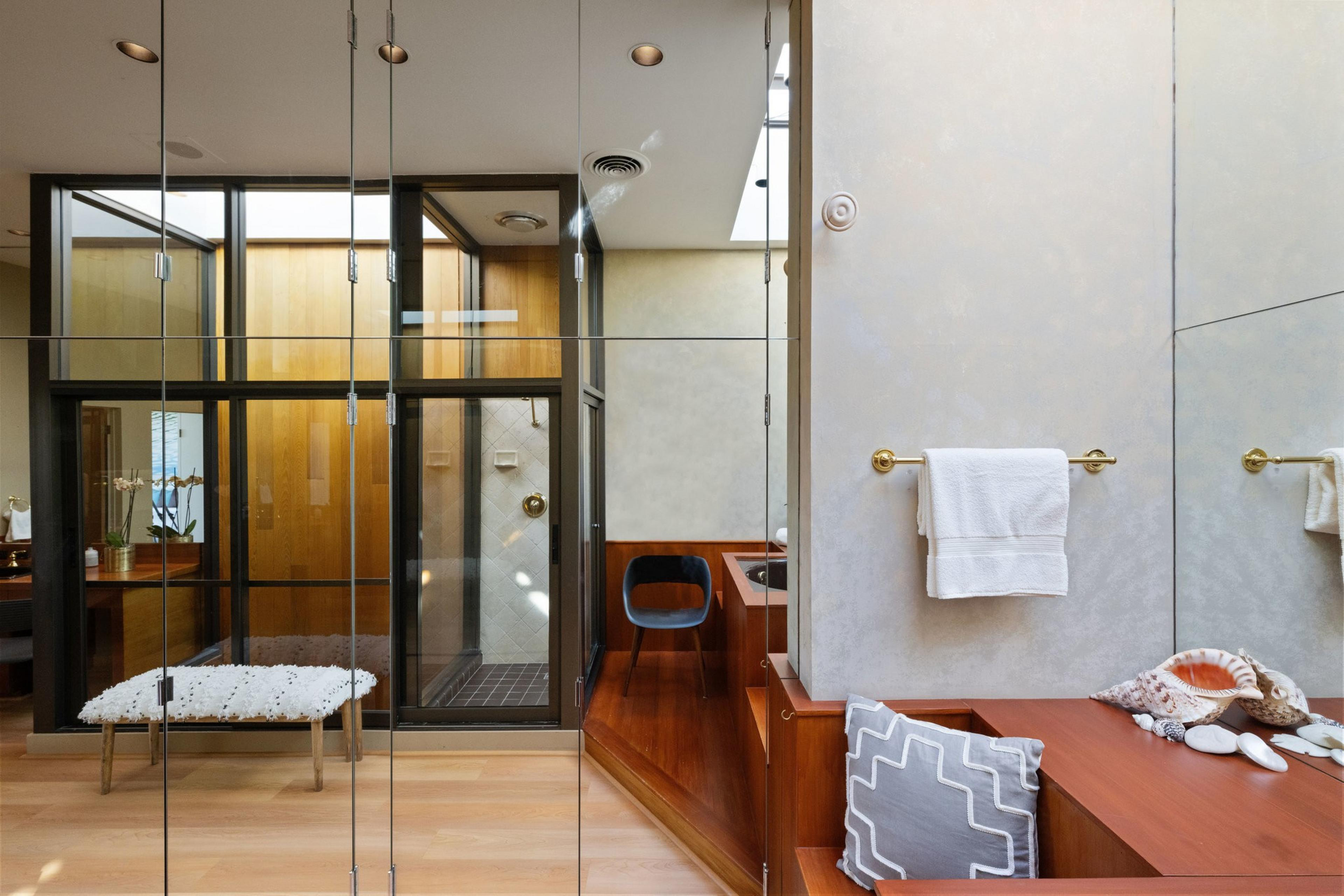 This image shows a modern bathroom with a wooden theme, a glass shower, a bench with a cushion, a towel on a gold holder, and seashells on a wooden surface.