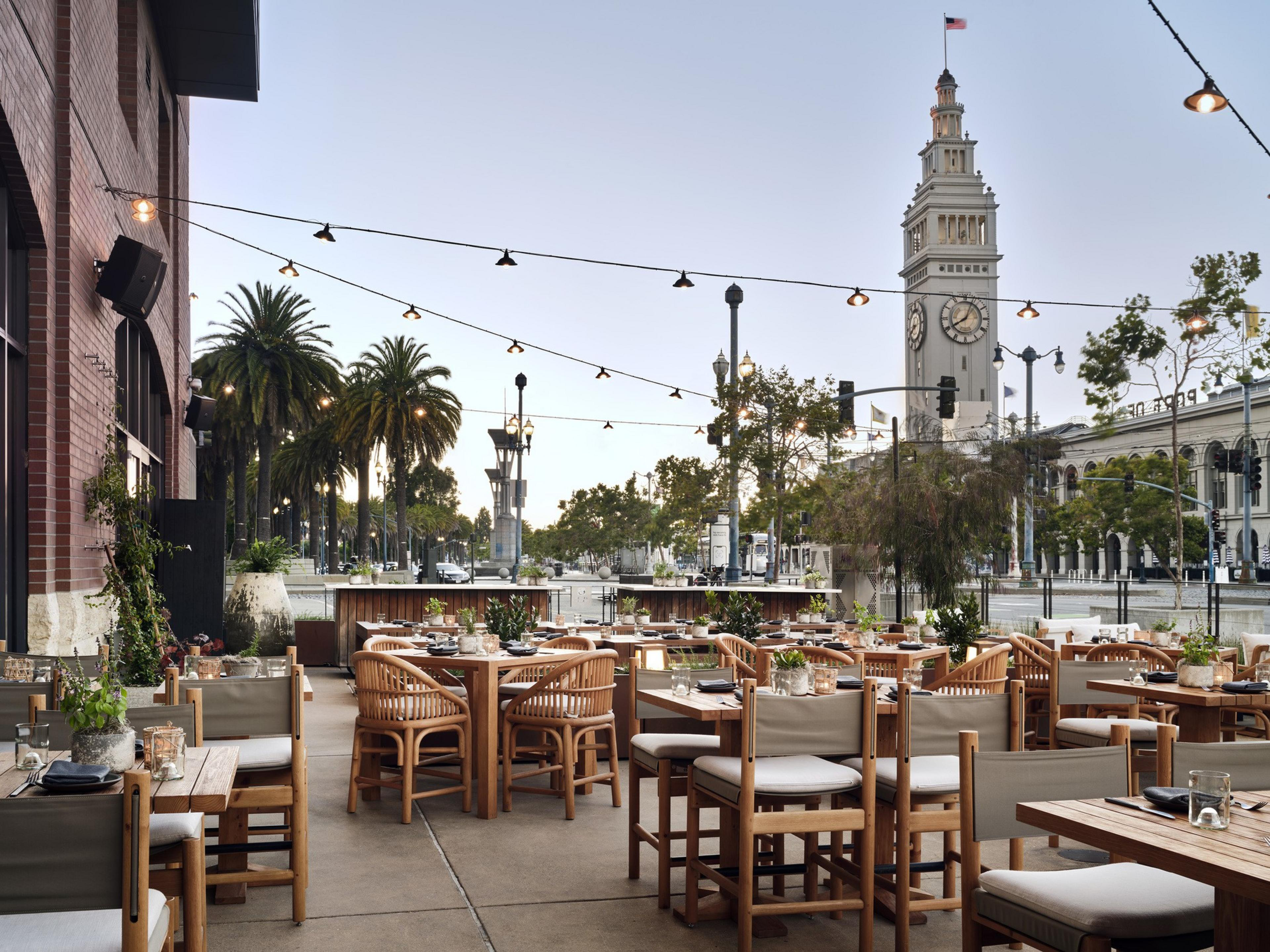 An outdoor restaurant patio with wooden tables and chairs, string lights overhead, palm trees, and a clock tower with an American flag in the background.