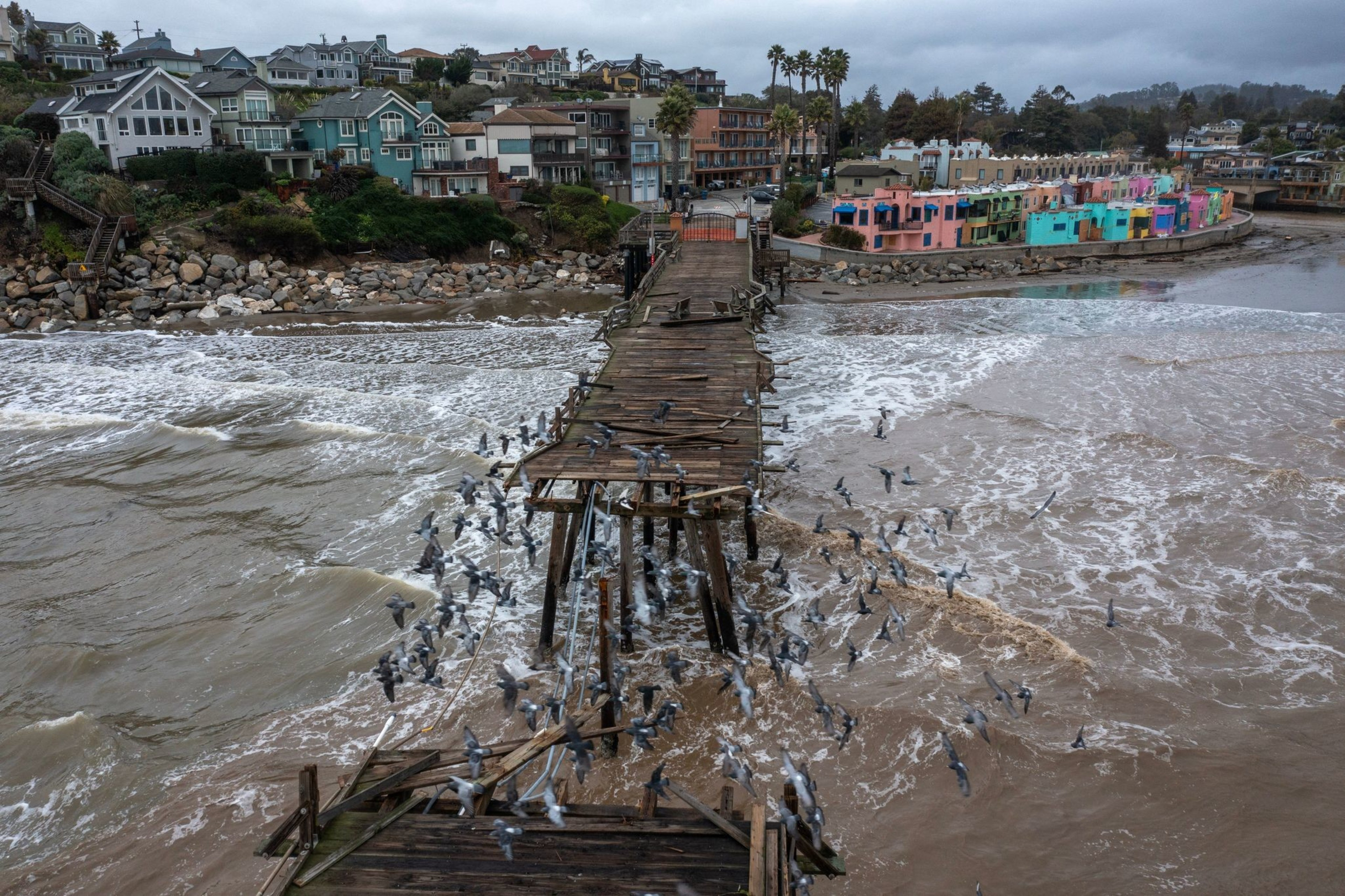 An aerial view of a birds flying over a wooden pier with a large gap seen next to colorful homes along the beach