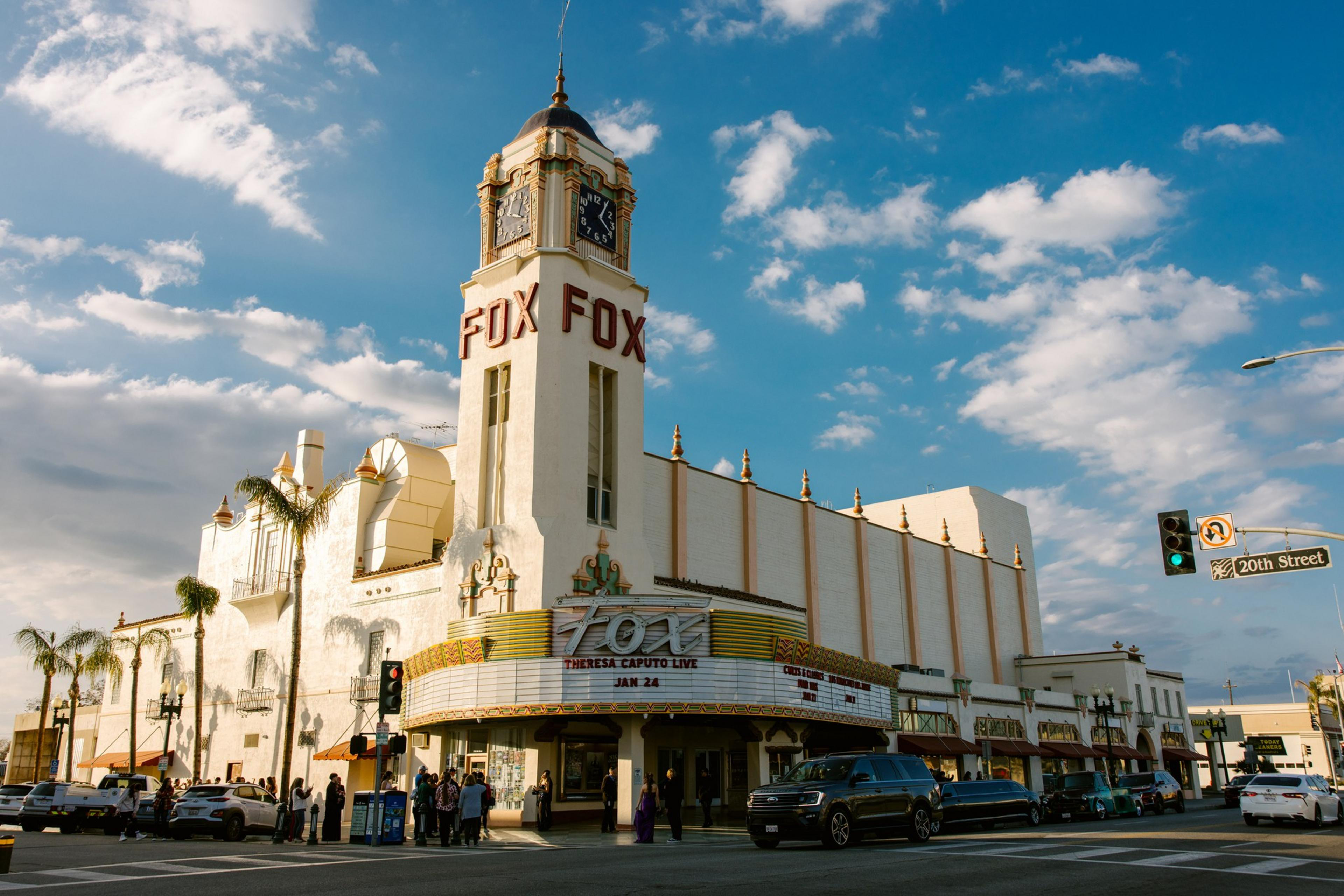 The image shows a historic theater with a tall tower labeled &quot;FOX.&quot; Its marquee advertises an event. Several people and cars are on the street under a bright sky.