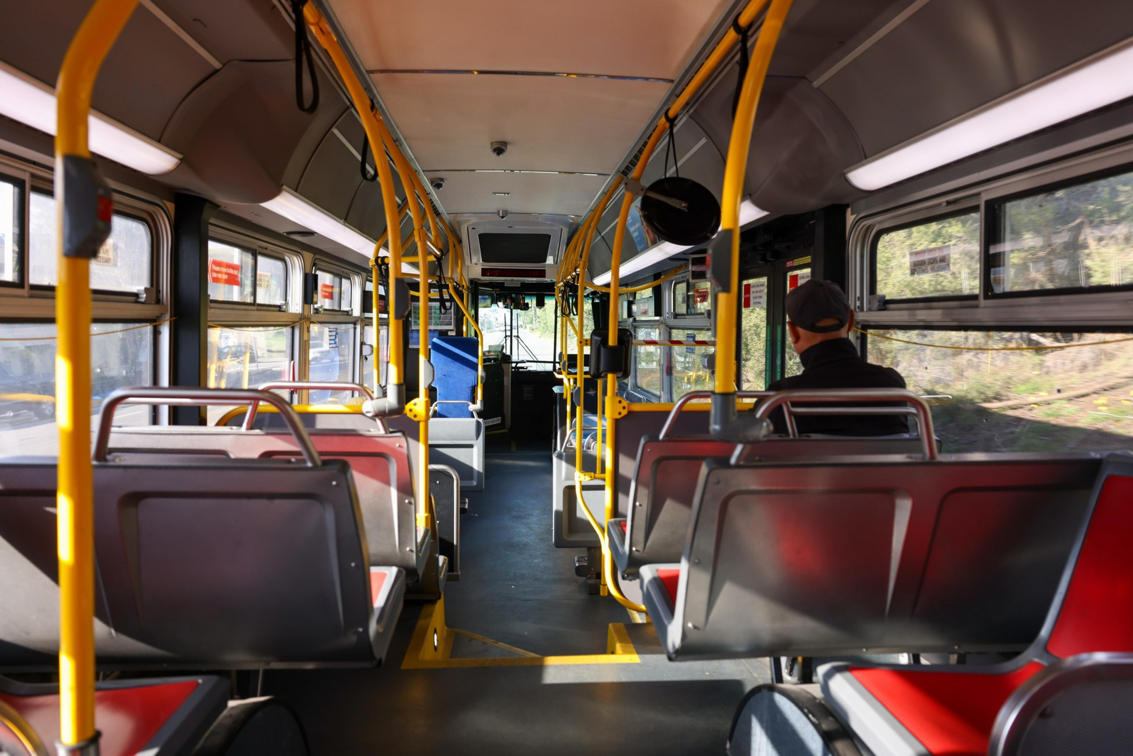 The image shows the inside of a nearly empty bus with yellow poles, red and gray seats, and a single occupant sitting towards the front.