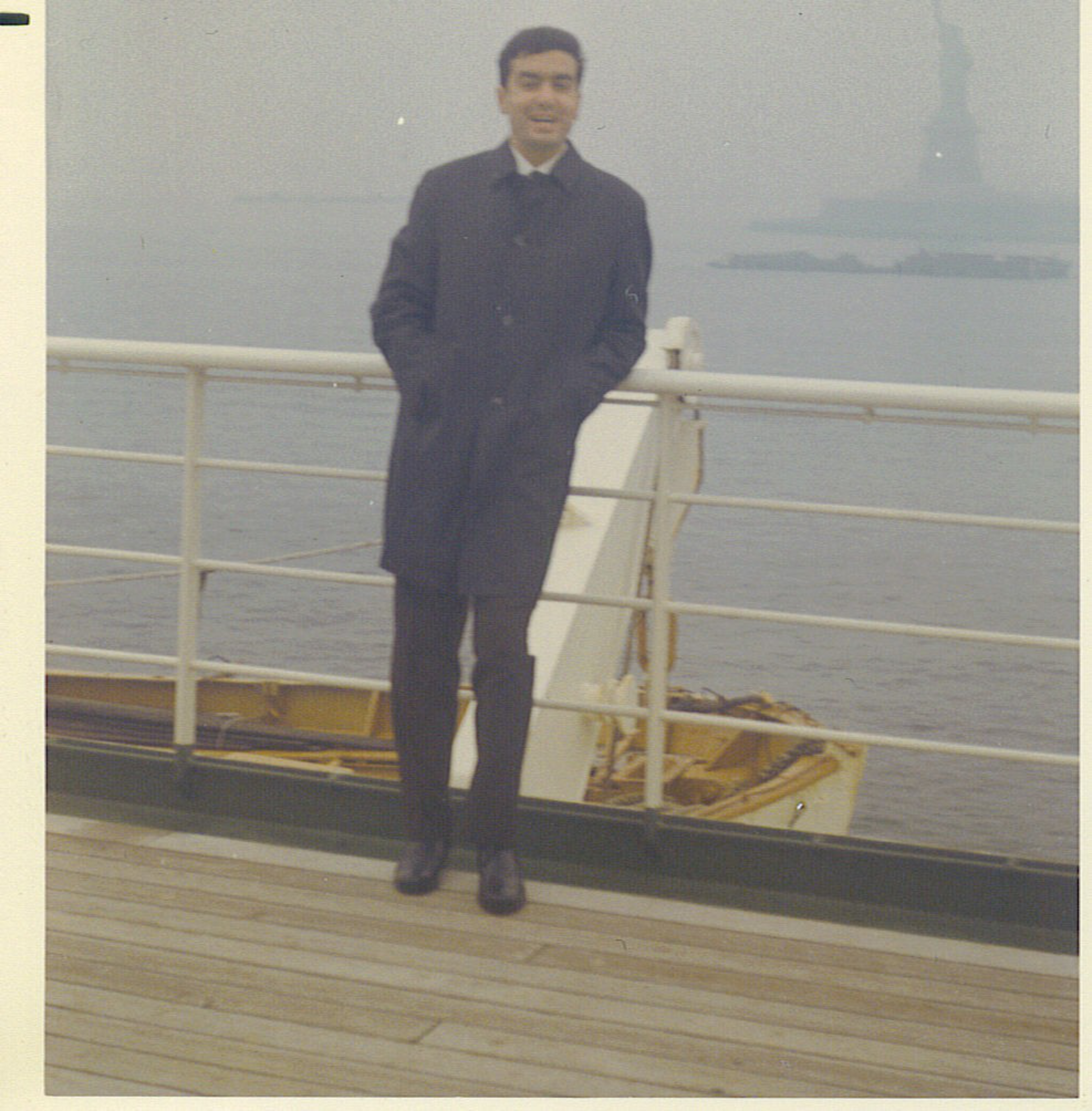 A man in a dark coat stands on a ship deck with hands in pockets, smiling, with the Statue of Liberty faintly visible in the foggy background across the water.