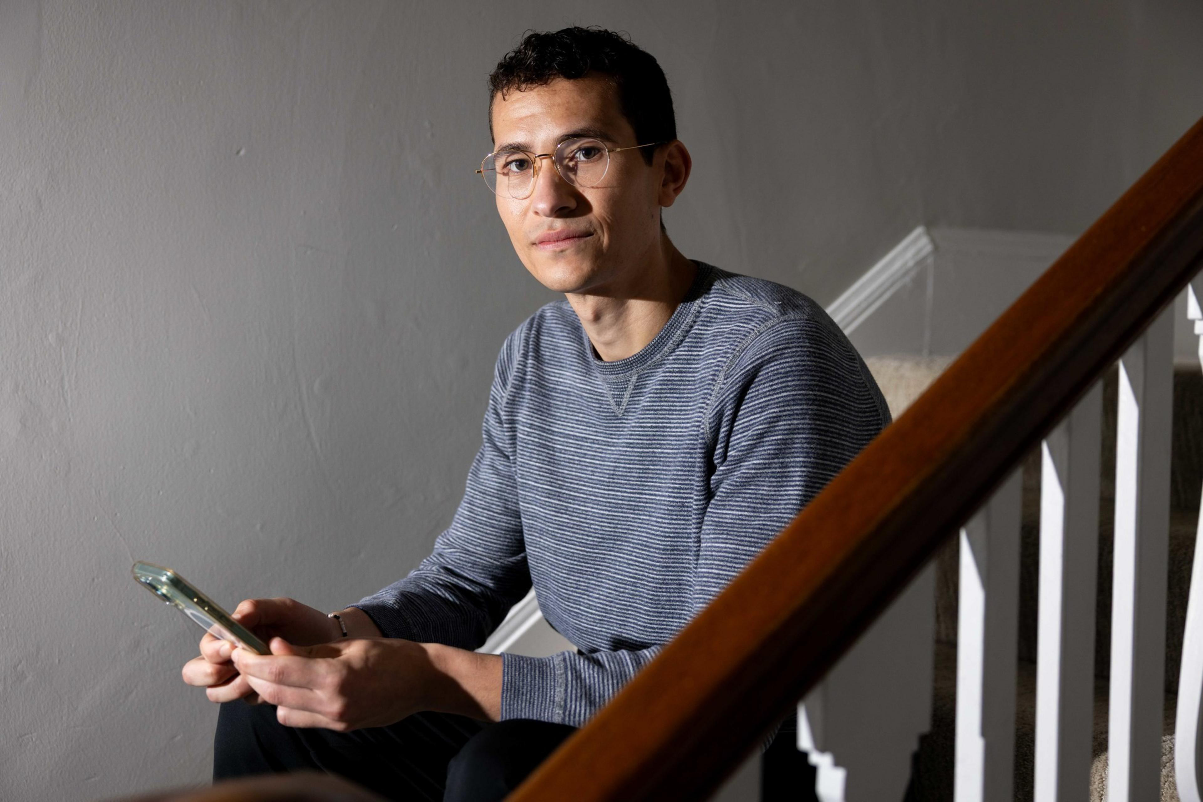A person wearing a striped sweater and glasses sits on a staircase, holding a smartphone, against a plain gray wall.