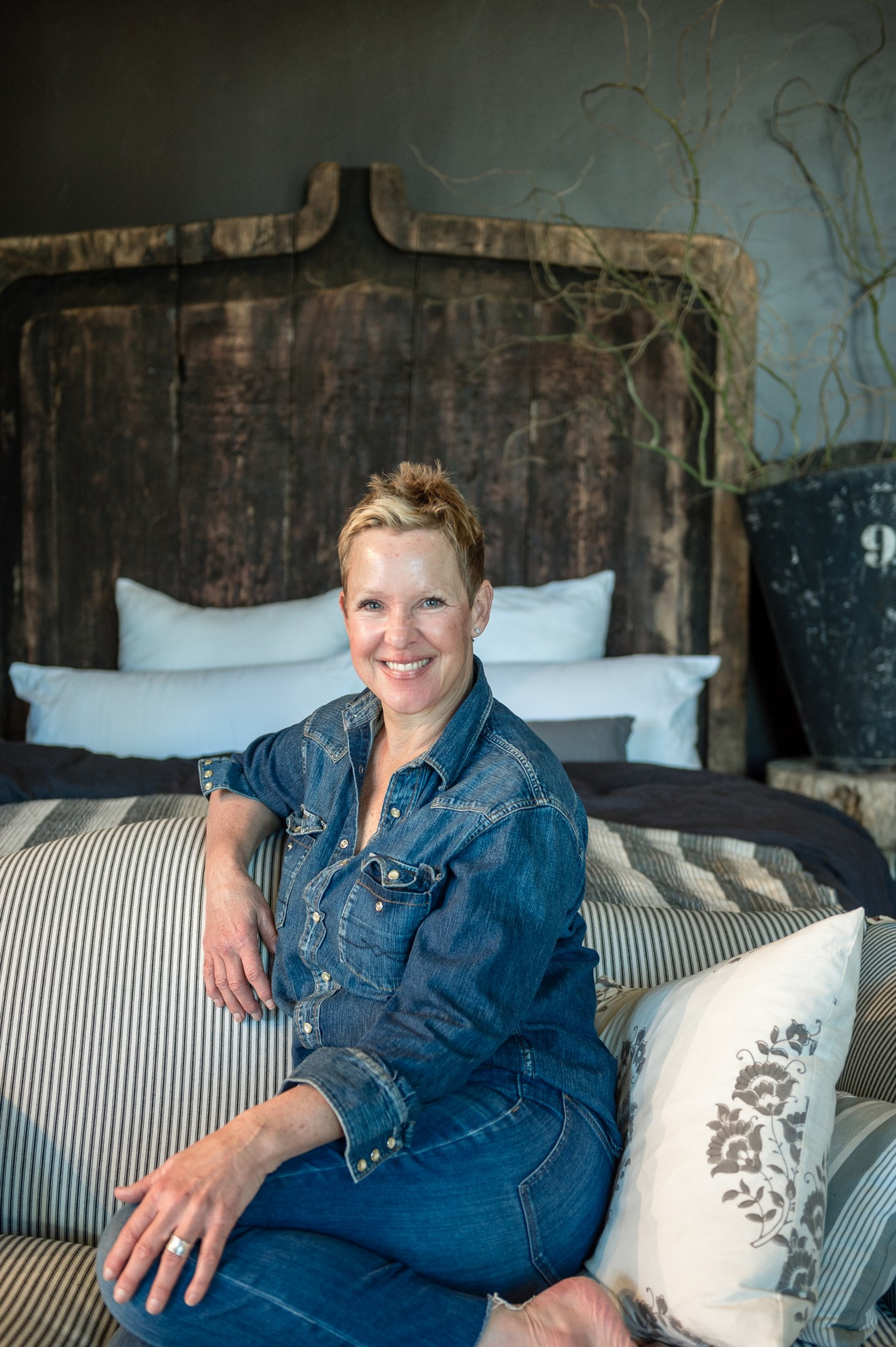 A smiling woman with short hair, wearing a denim shirt and jeans, sits barefoot on a striped couch with a floral pillow in a cozy room.