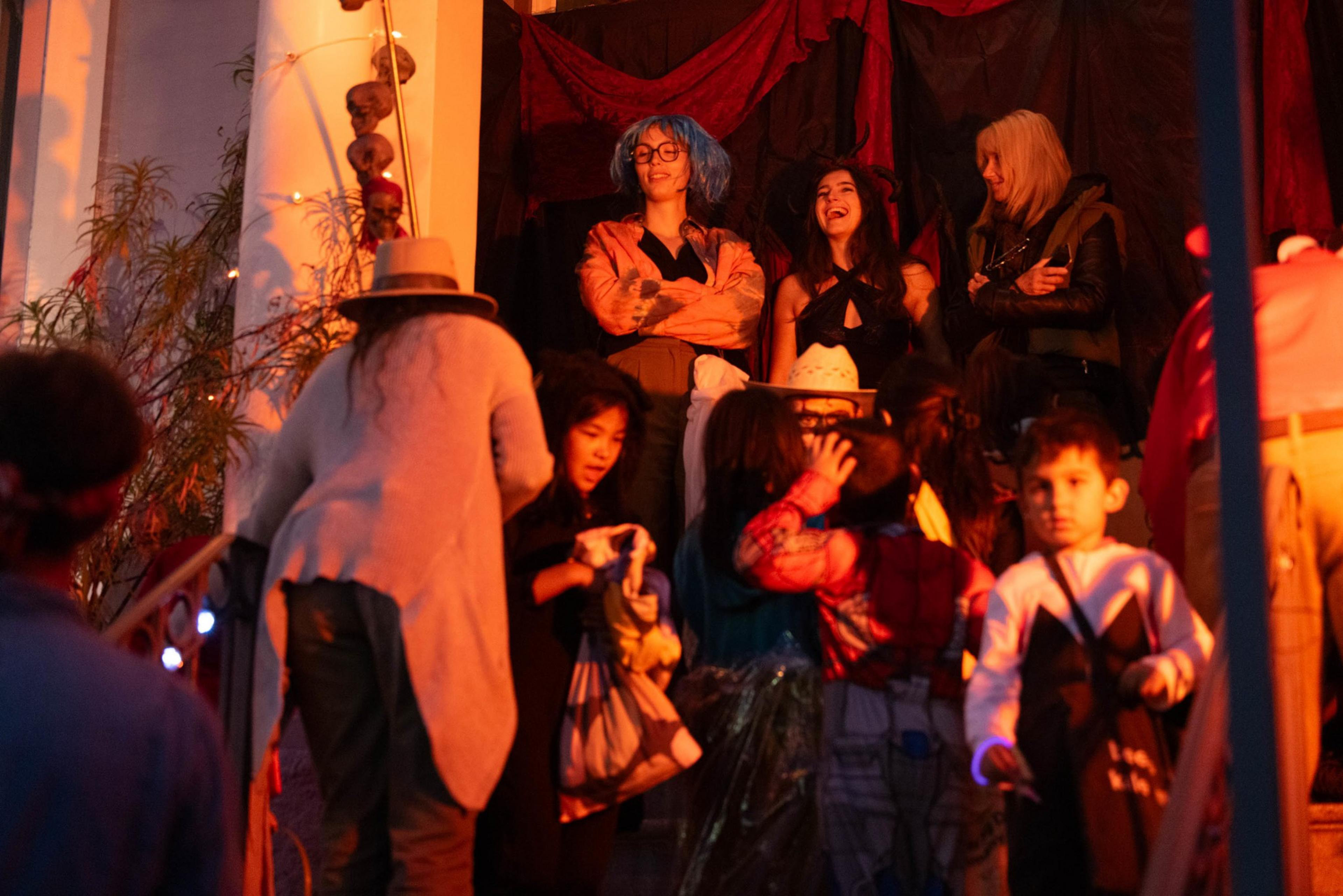 A group of children and adults gather on and around a porch lit with warm orange light, with three women standing and interacting at the top.