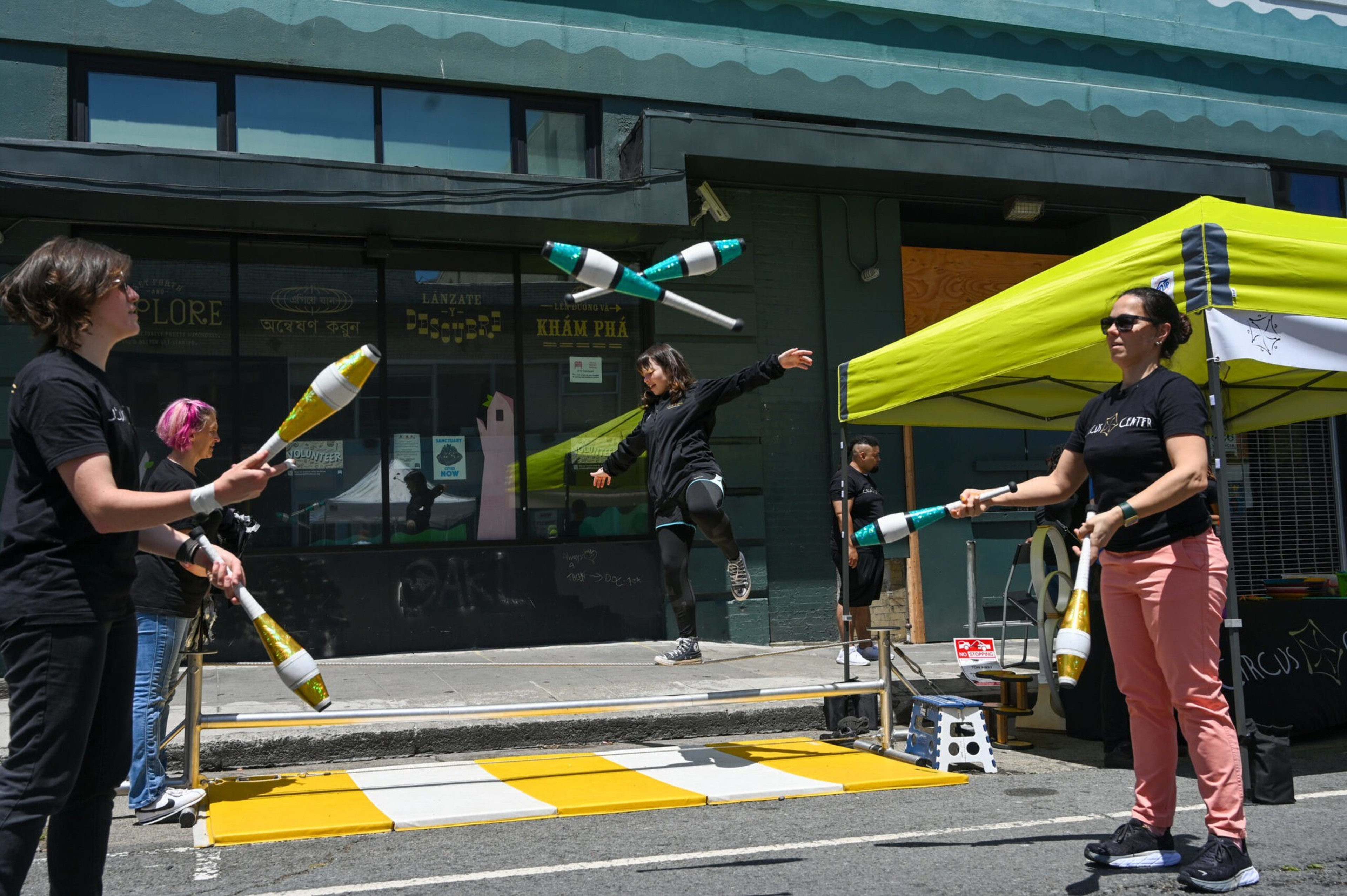 Two people are juggling clubs on a street. In the background, someone crosses a tightrope.