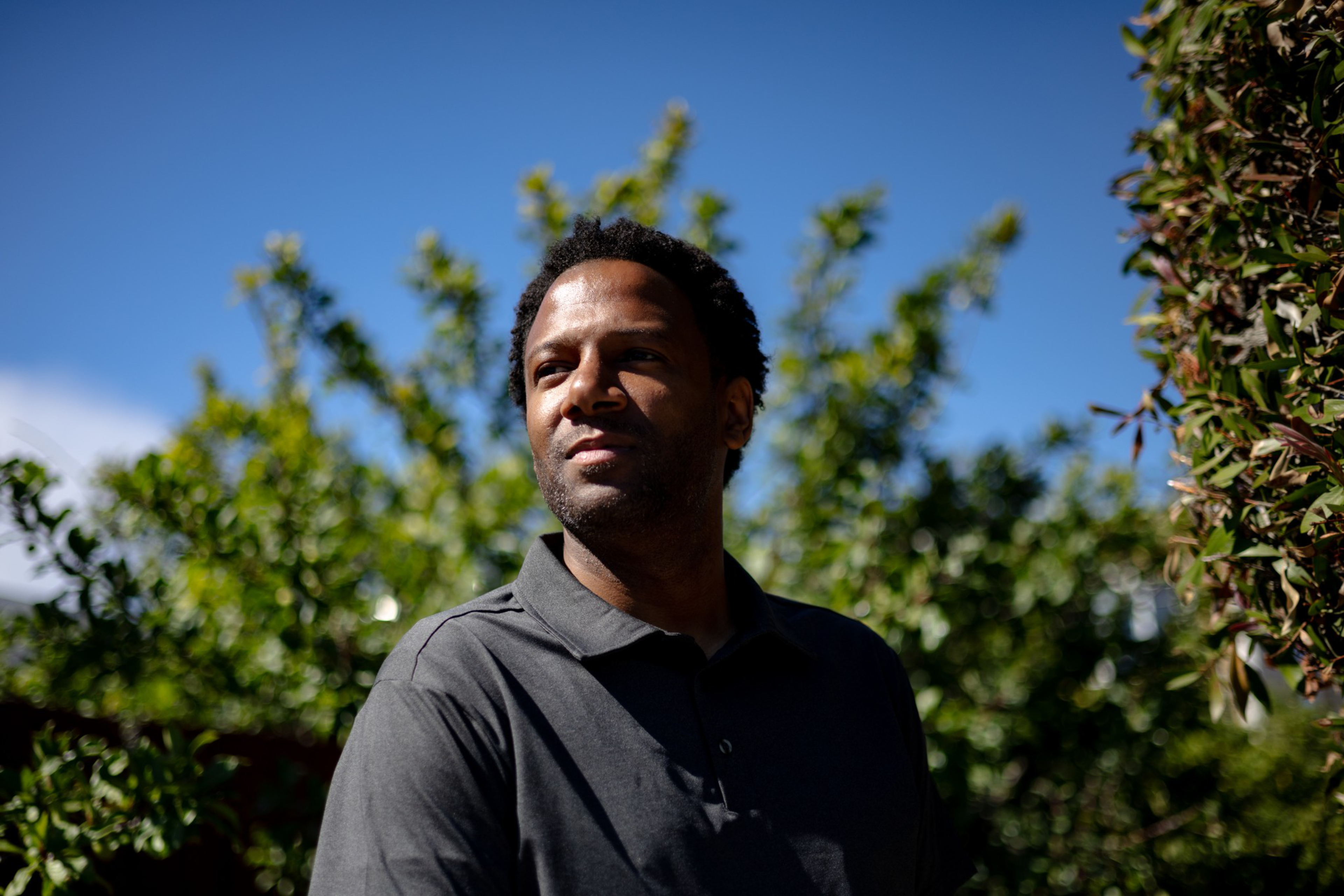 A man wearing a dark polo shirt stands outdoors, looking slightly to his left, with green leafy trees and a clear blue sky behind him.