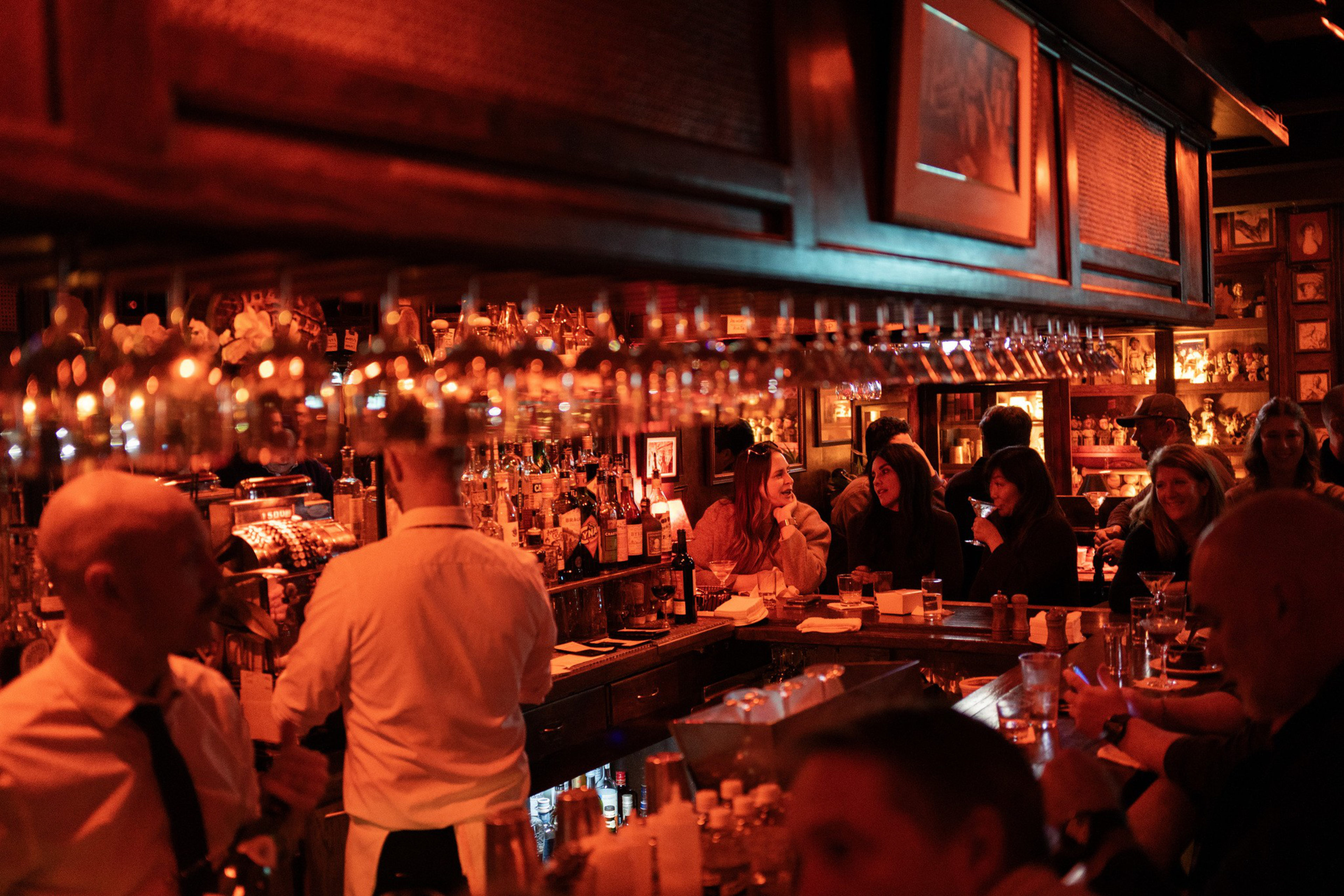 A warmly lit bar with patrons chatting and bartenders serving drinks, surrounded by shelves of bottles and hanging glassware overhead.