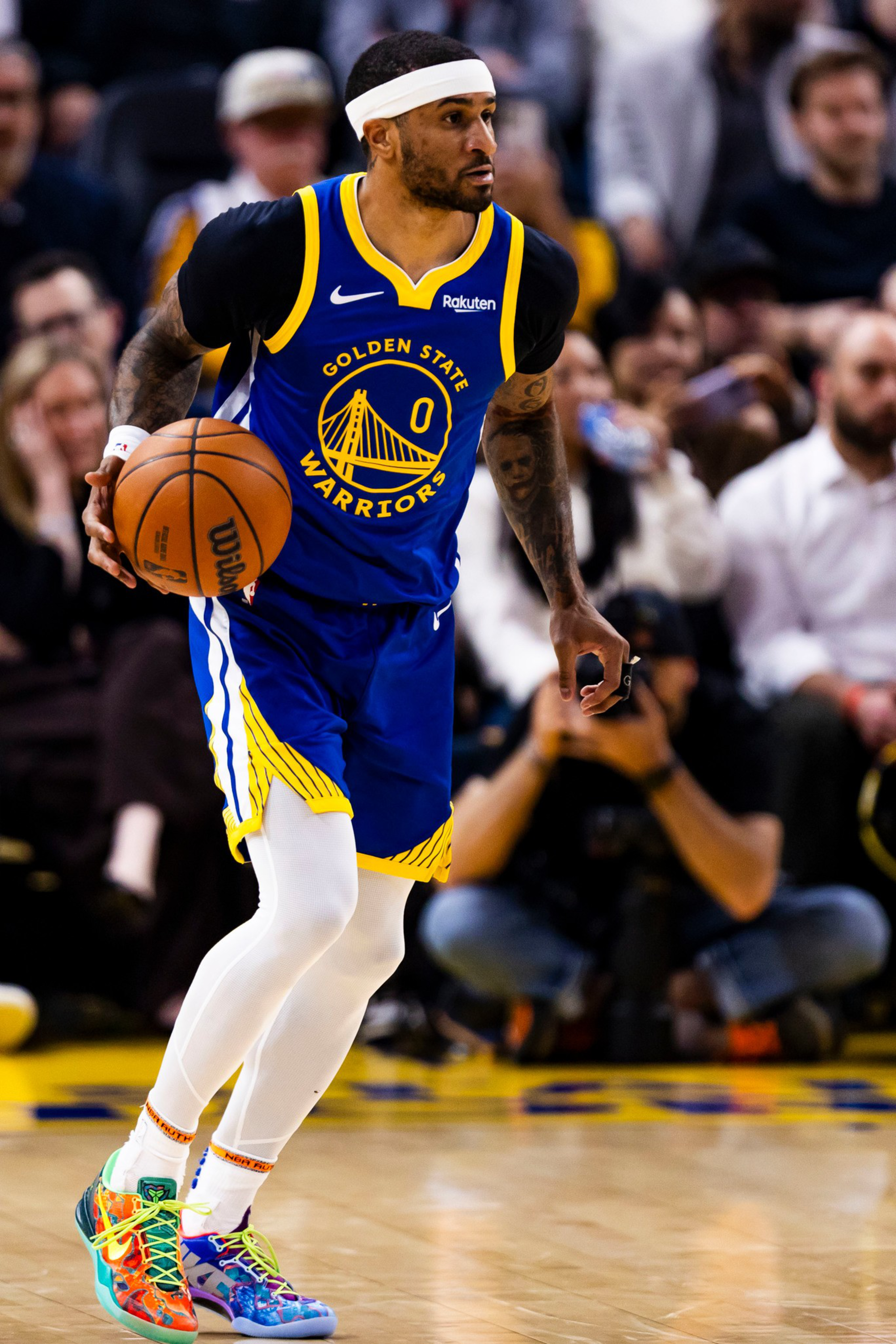 A Golden State Warriors player in a blue and yellow uniform dribbles a basketball on the court with spectators blurred in the background.