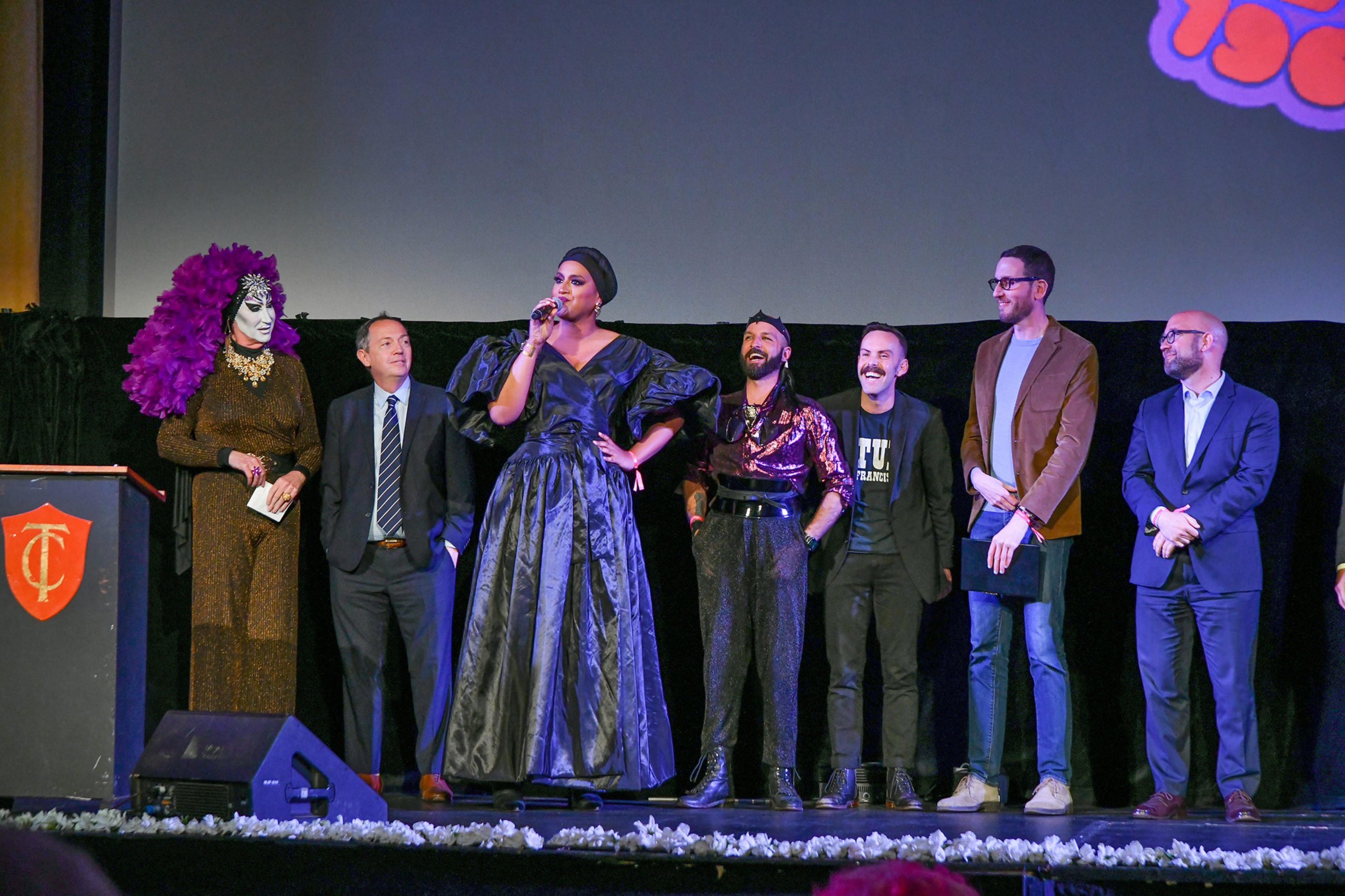 Seven people stand on a stage, with one person in a dramatic black gown speaking into a microphone and others dressed in formal or stylish attire.