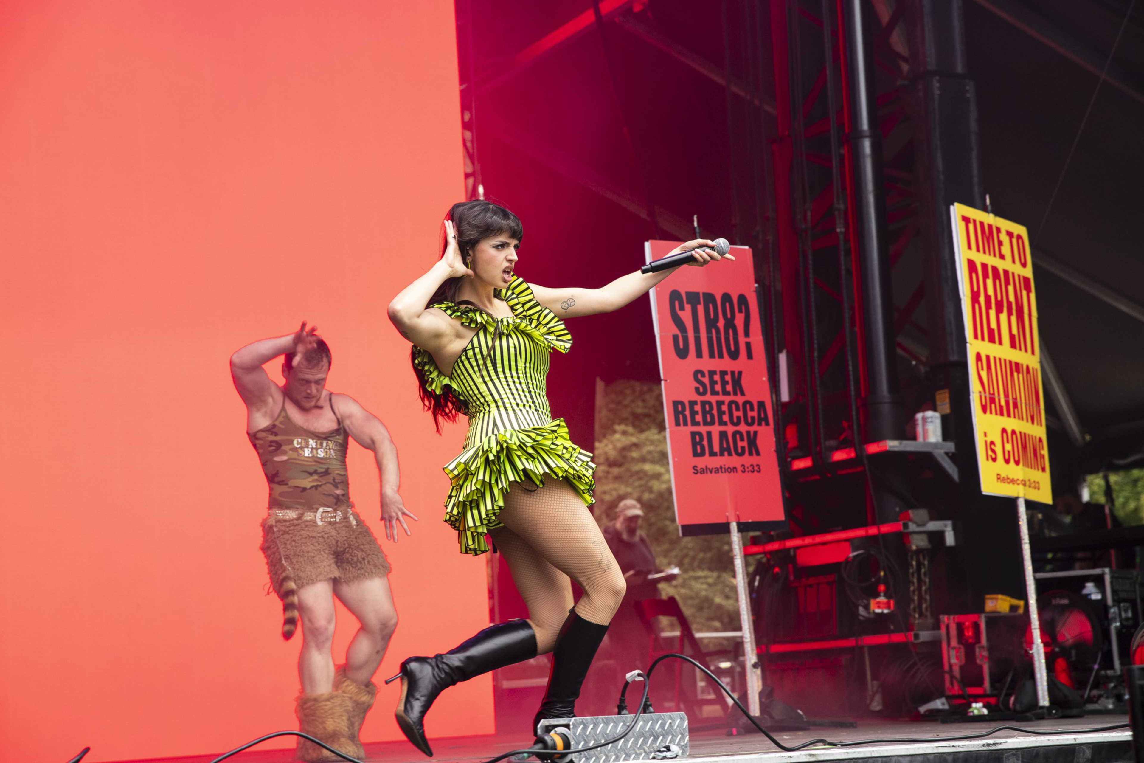 A female performer in a green striped dress and black boots dances on stage while a man in a furry outfit dances behind her, with red lighting and protest signs.