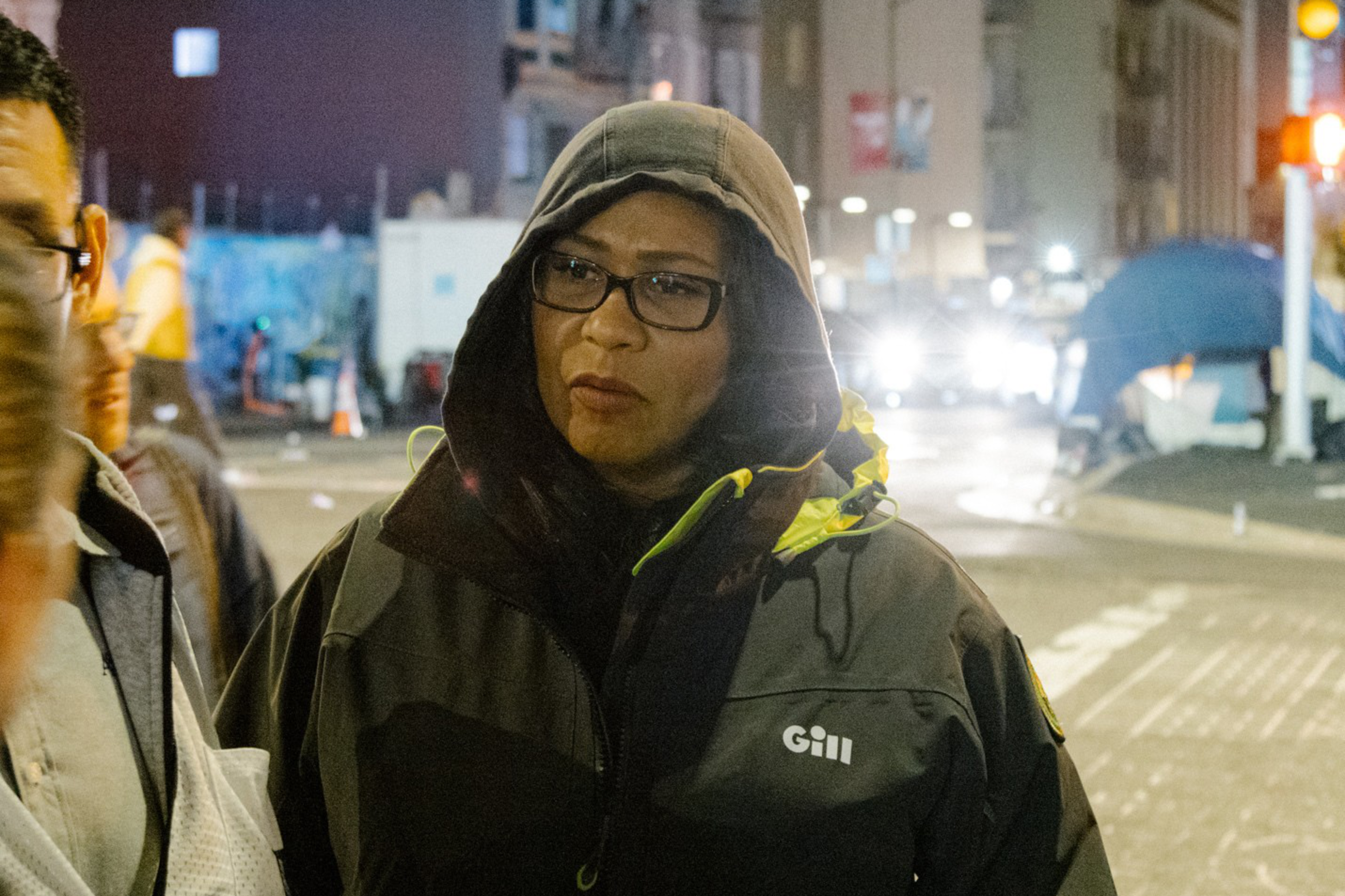 A woman in a hooded jacket stands on a bustling city street at night, with illuminated buildings and tents in the background.