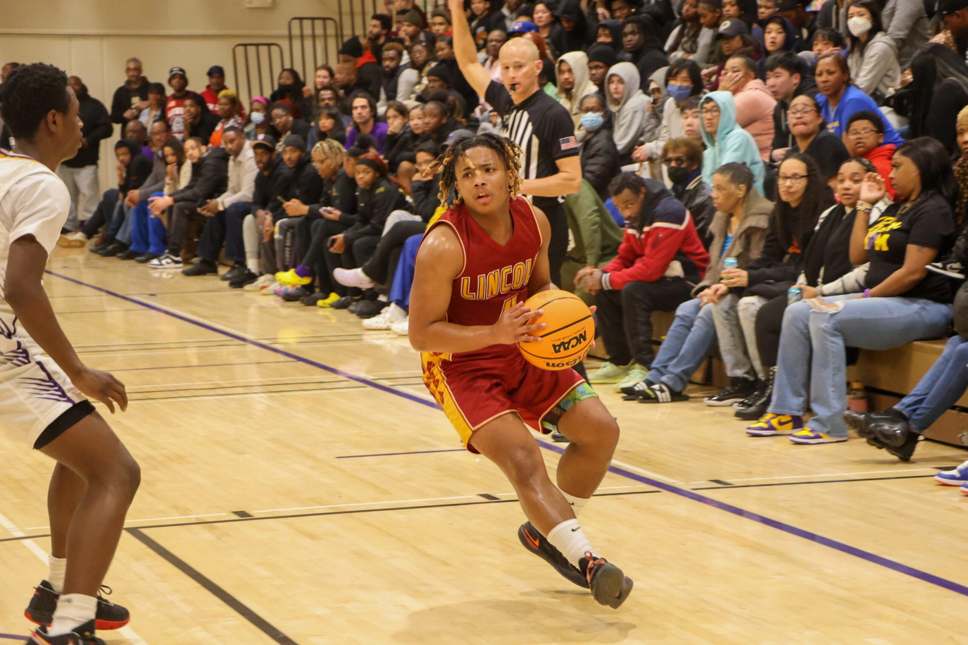 A basketball player in a red "LINCOLN" uniform maneuvers with the ball as a defender approaches, while spectators watch intently from the bleachers.