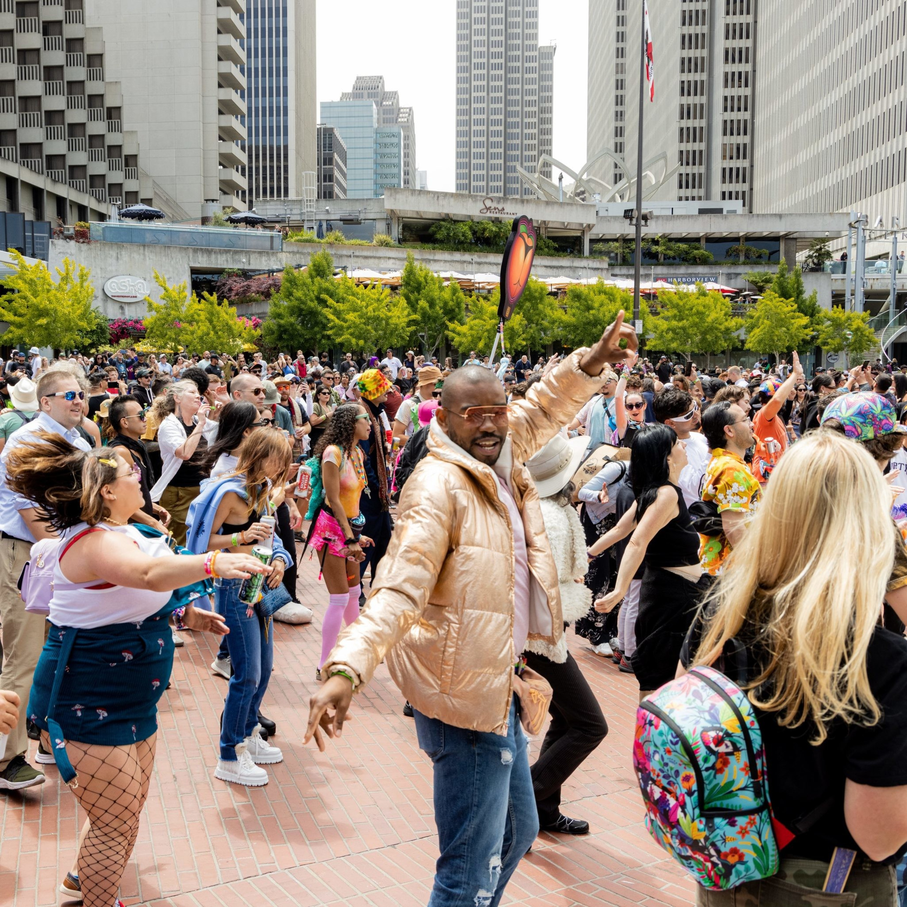 A joyful crowd dances at an outdoor city event surrounded by tall buildings, vibrant trees, and lively people in colorful outfits, exuding energy and fun.