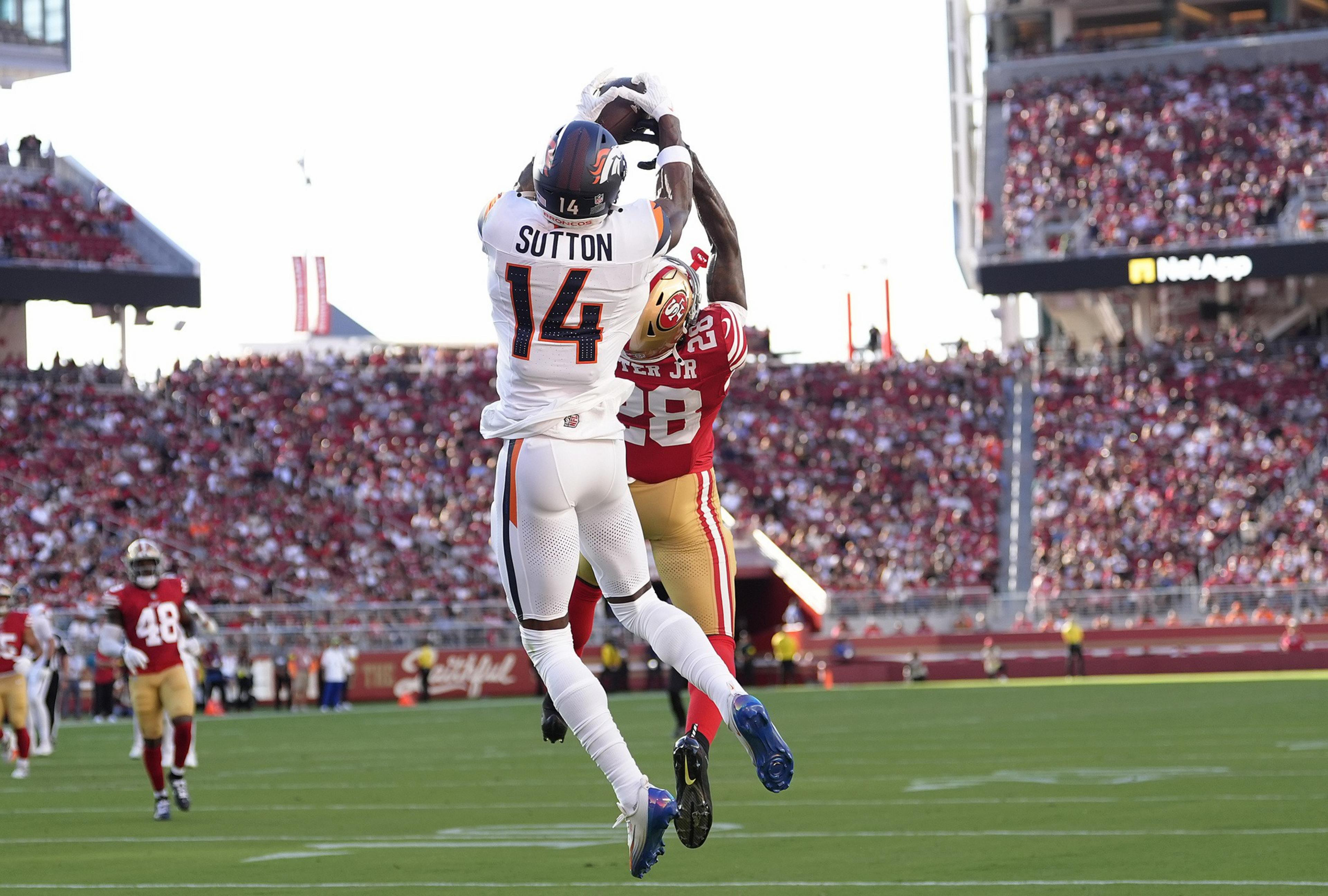 A player in white jersey number 14 leaps to catch a football while a player in a red jersey number 28 defends during a crowded stadium game.