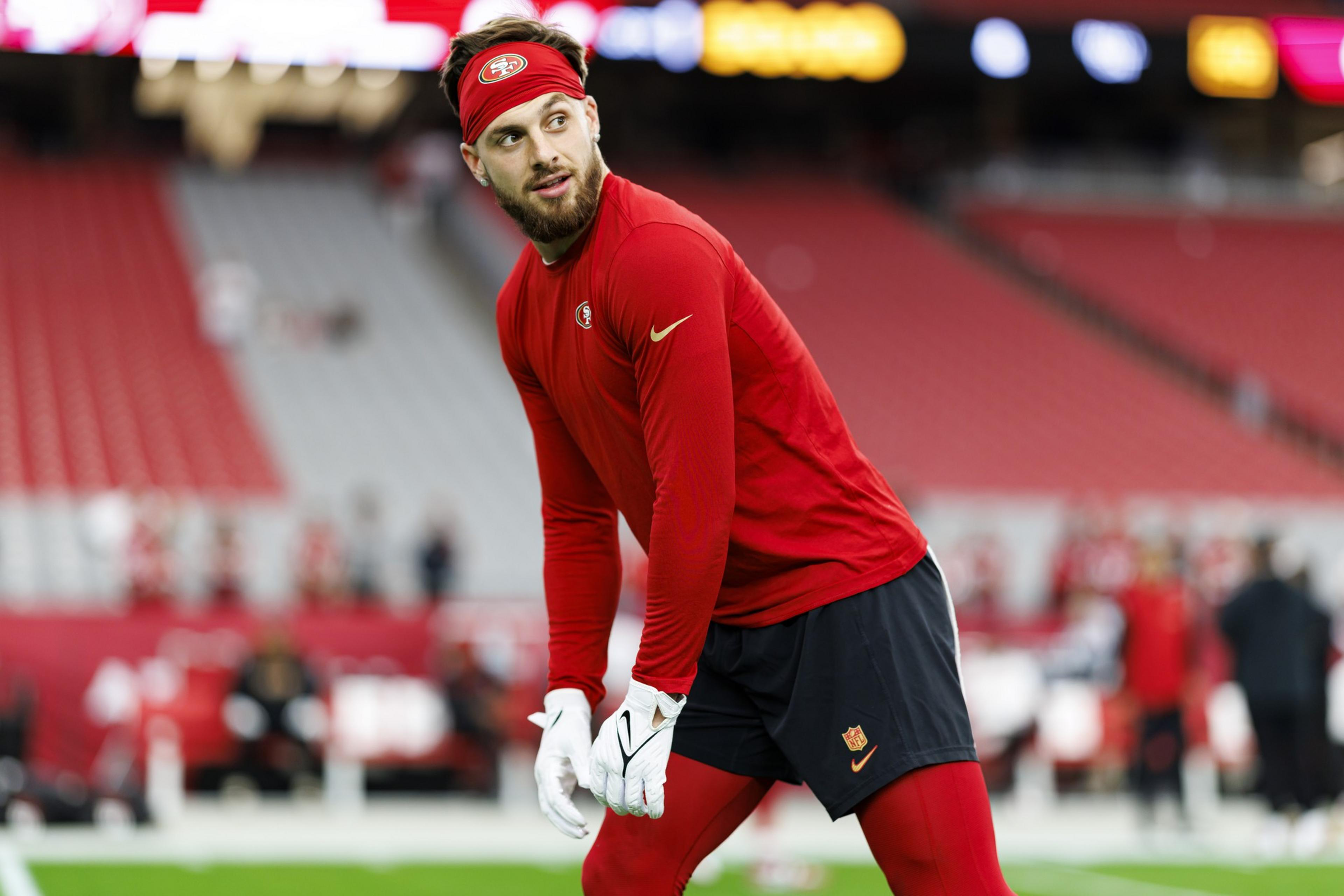 A man in a red long-sleeve shirt, black shorts, red leggings, white gloves, and a red headband is preparing on a football field.