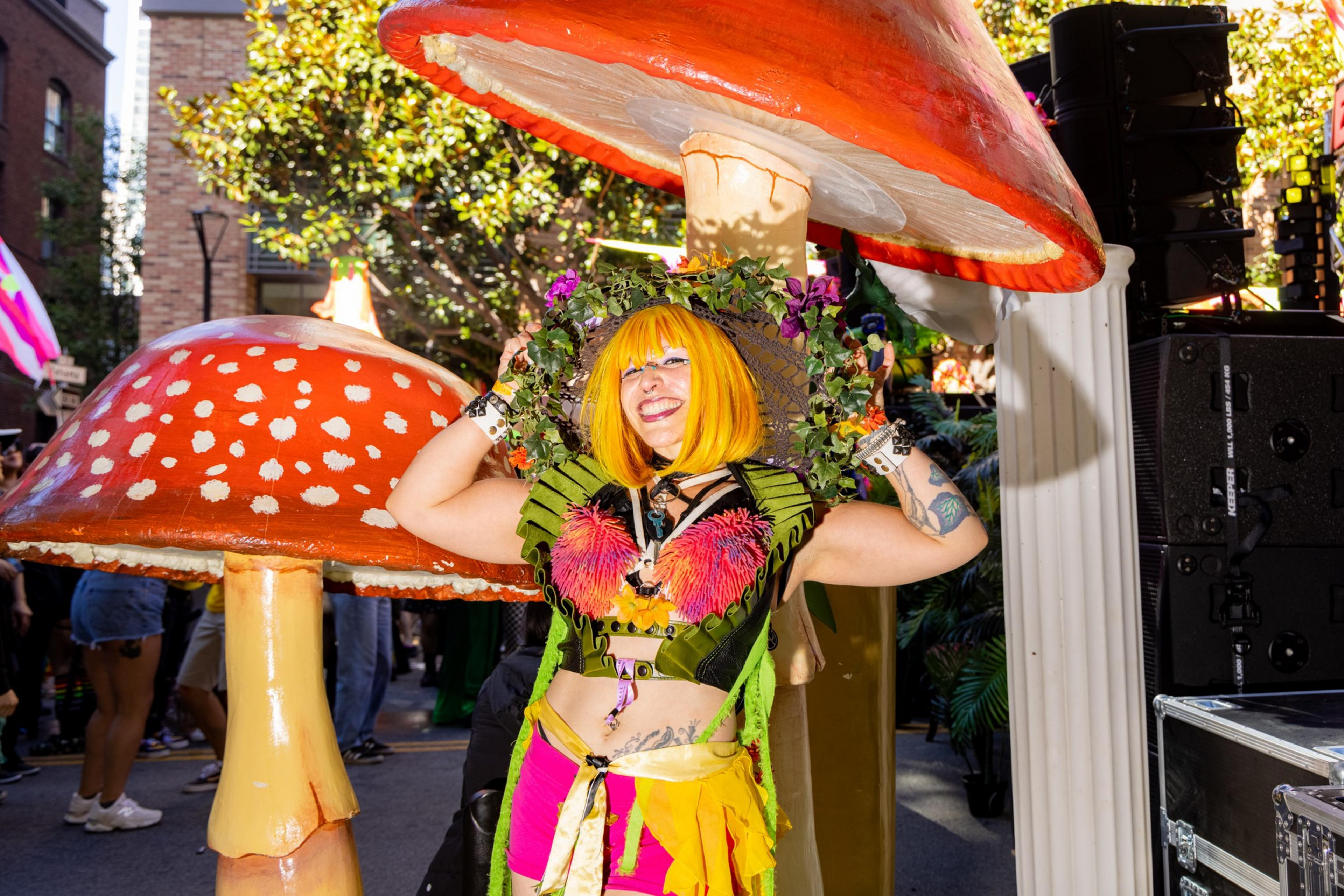 A person with bright yellow hair and colorful, eclectic clothing poses cheerfully between two large, whimsical mushroom decorations in an outdoor setting.