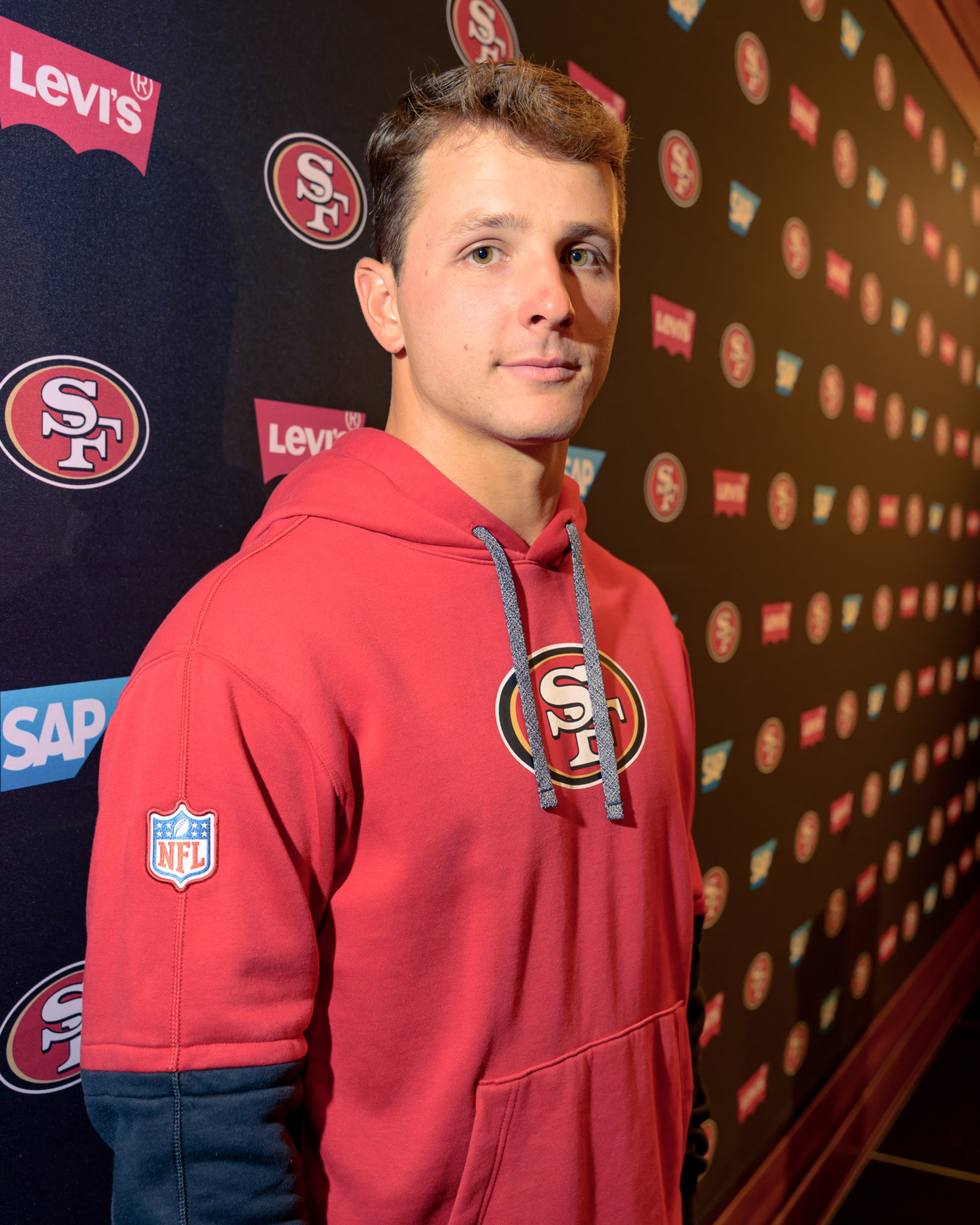 A person in a red hoodie with an "SF" logo stands against a backdrop featuring repeated logos of Levi's, SAP, and the same "SF" design.