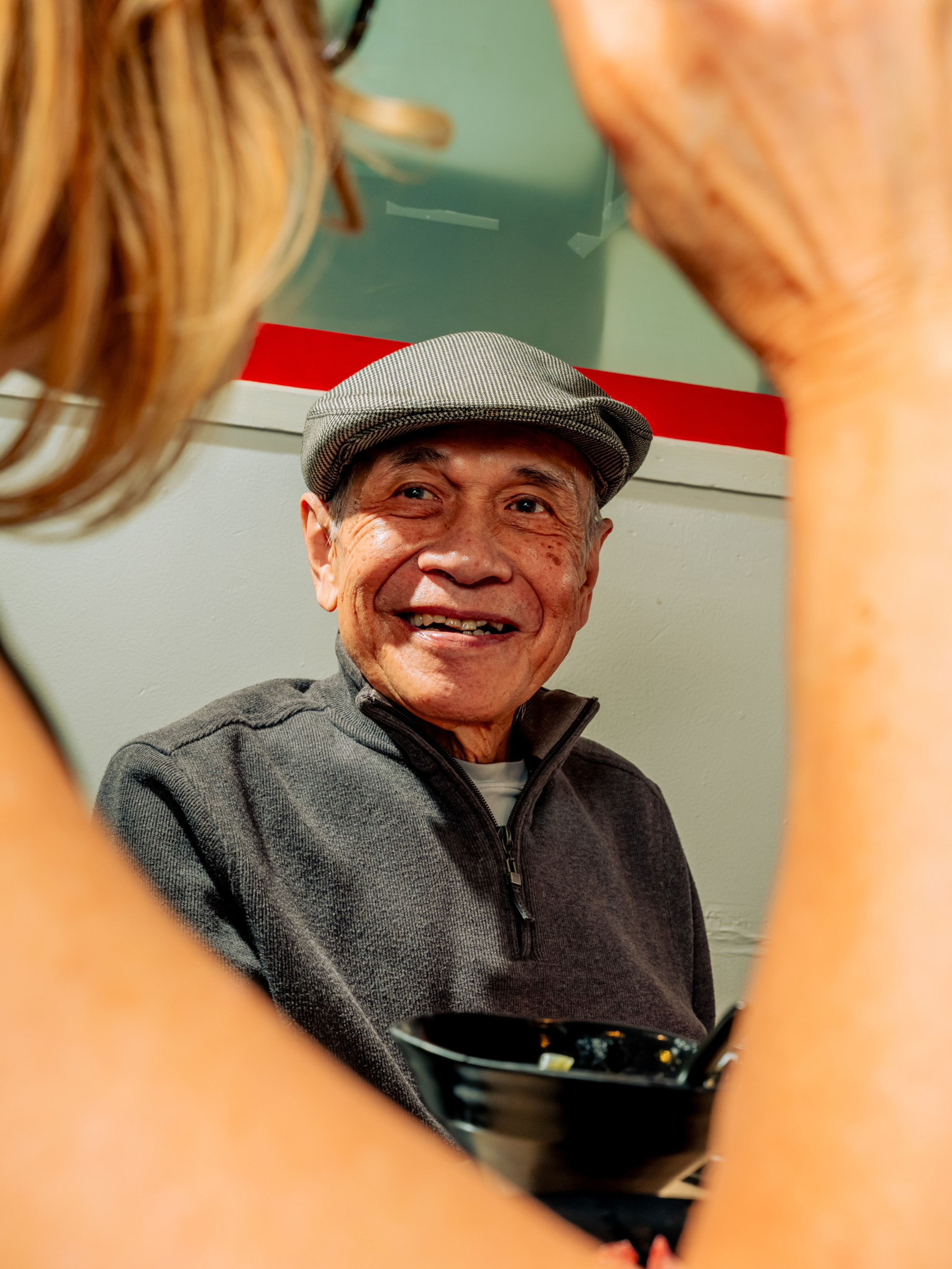 An older man wearing a gray flat cap and zip-up sweater smiles warmly while sitting at a table with a black bowl in front of him.