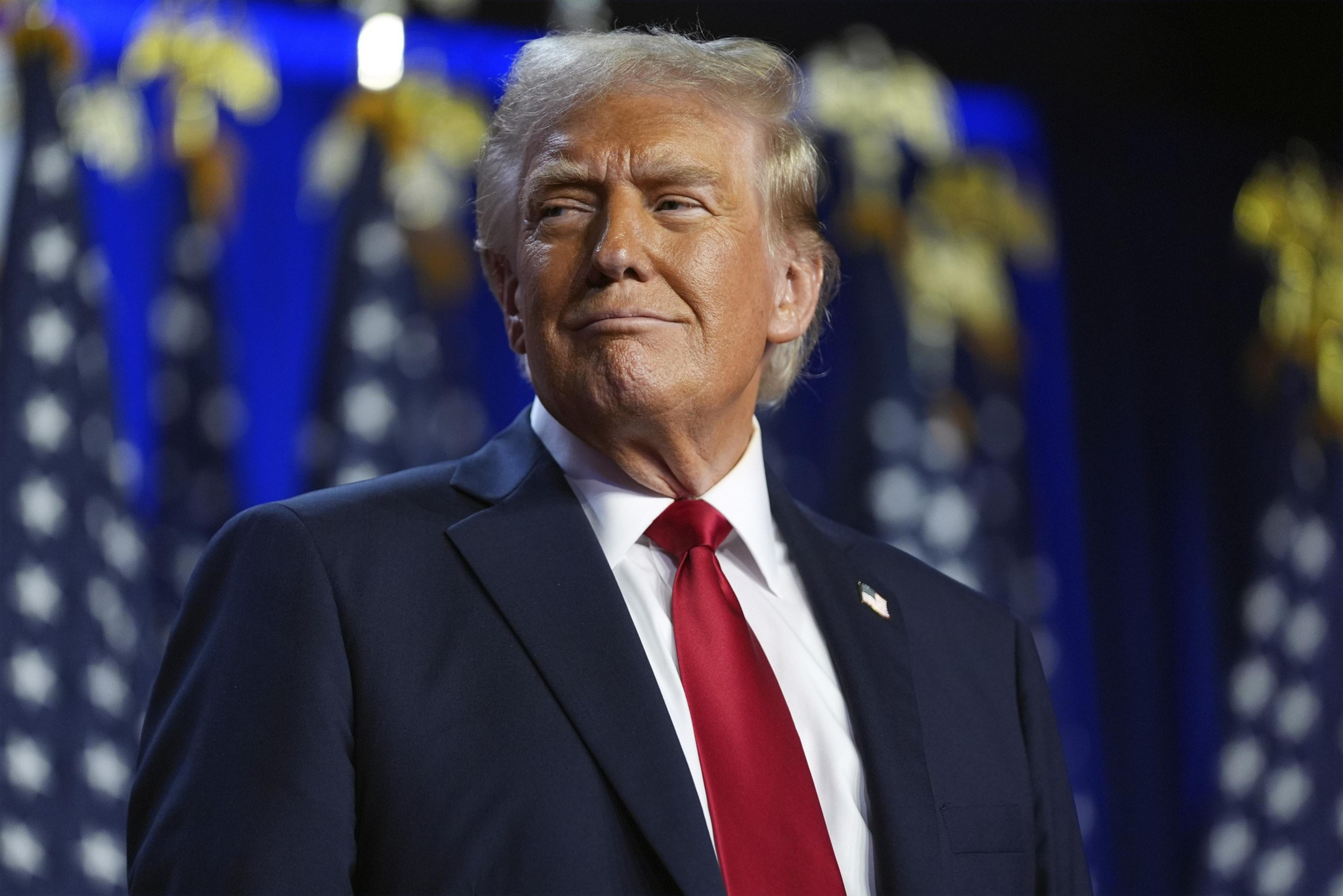 A person with blond hair is wearing a dark suit and red tie, looking to the side with a confident expression. The background shows blurred US flags.