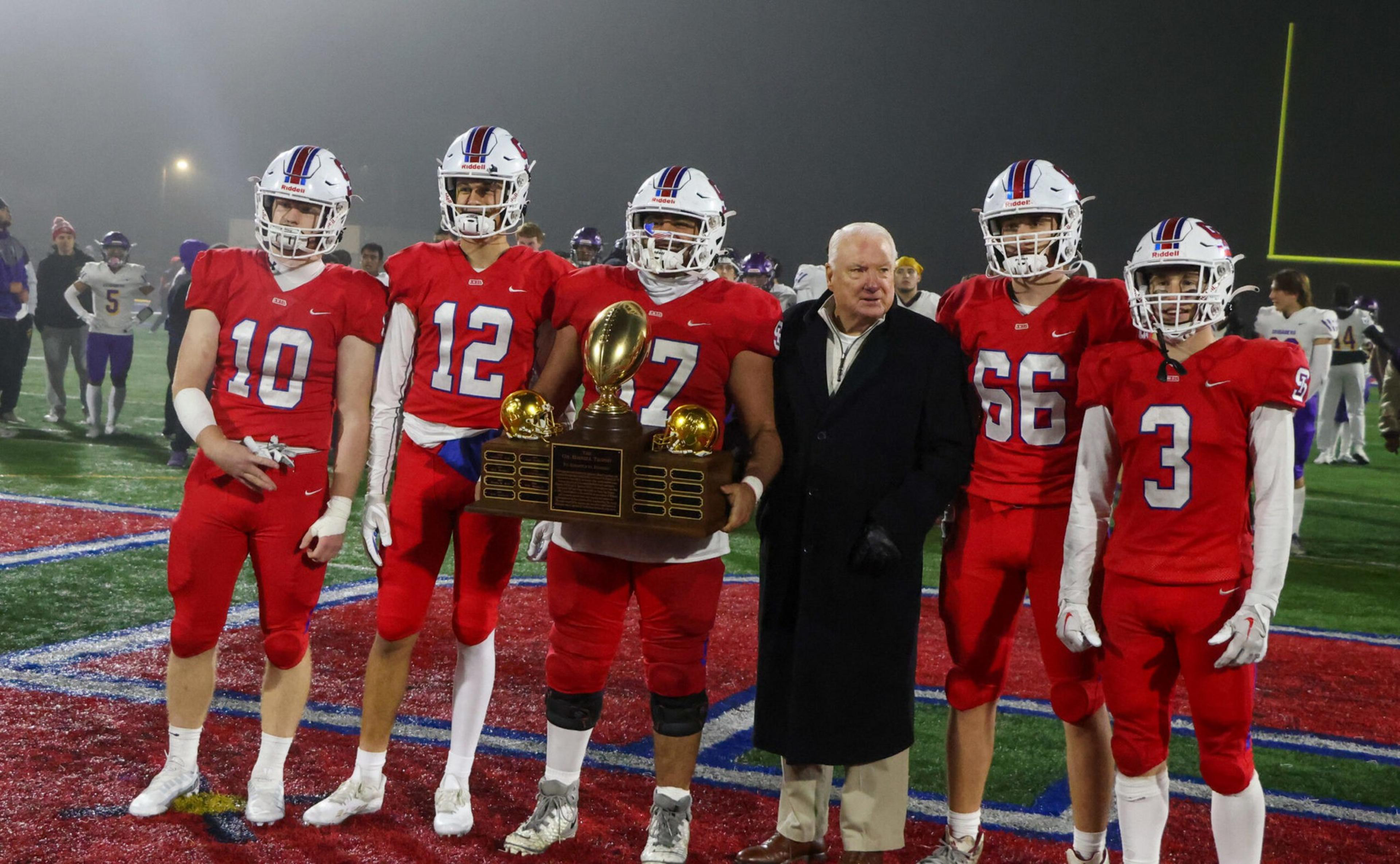 Five football players in red uniforms and one man in a black coat stand on a field, with the center player holding a large trophy.